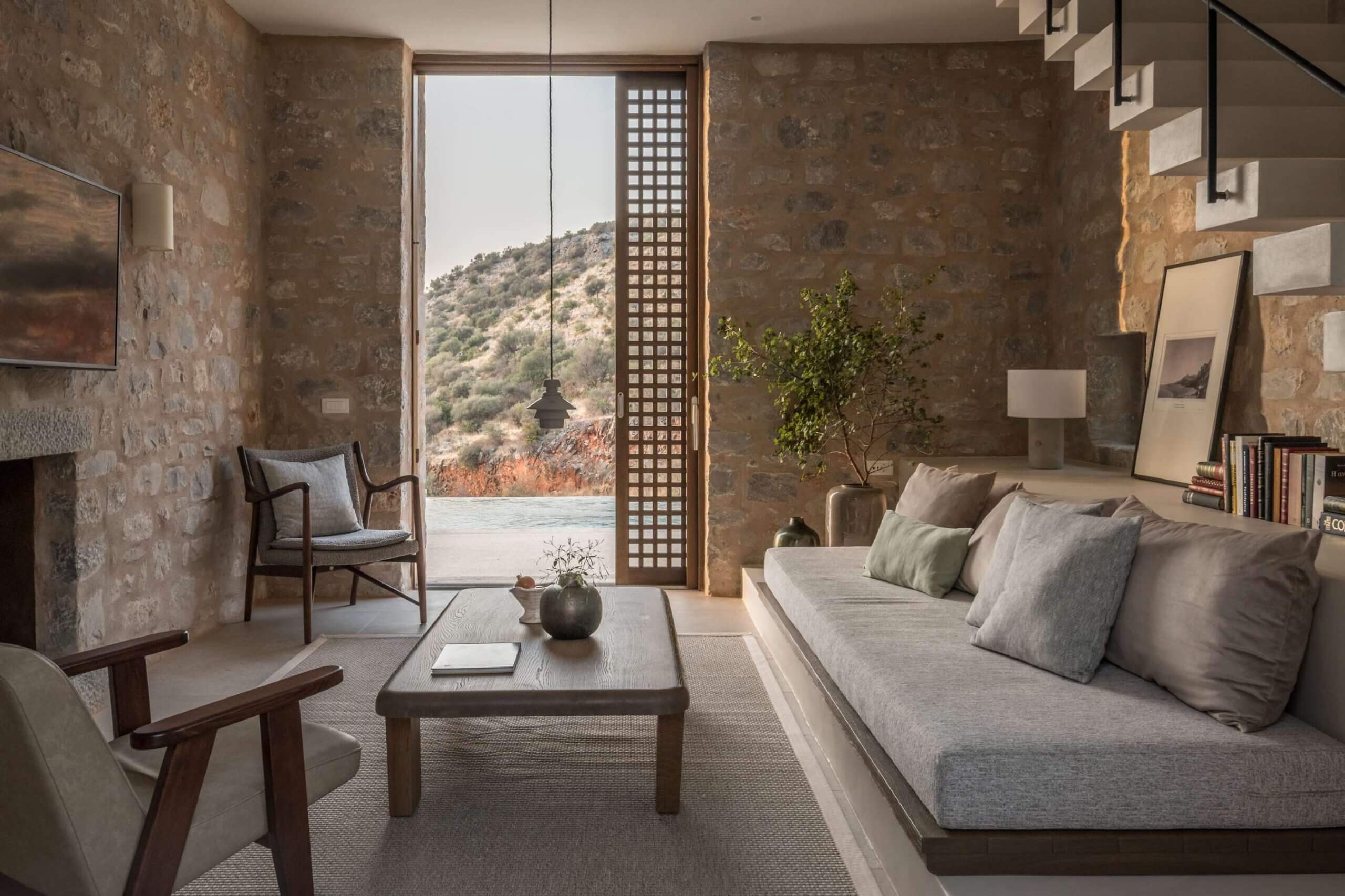 Monolithoi, Europe — Stone-walled bedroom with linen sofa, wooden furniture, mountain vista through floor-to-ceiling glazing, and minimalist Mediterranean interior design.