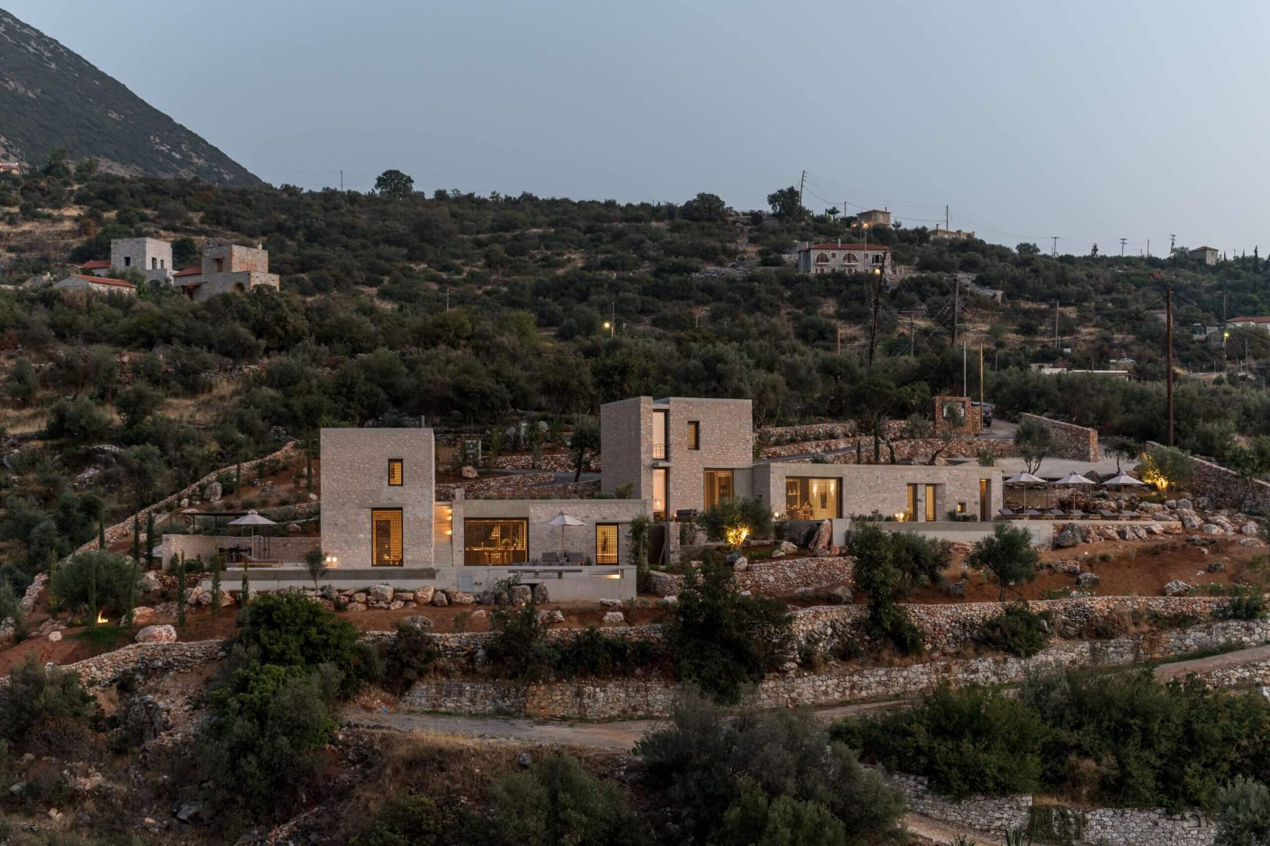 Monolithoi, Europe — Modern stone villa with illuminated windows nestled in terraced hillside landscape at dusk, surrounded by olive trees and limestone walls.