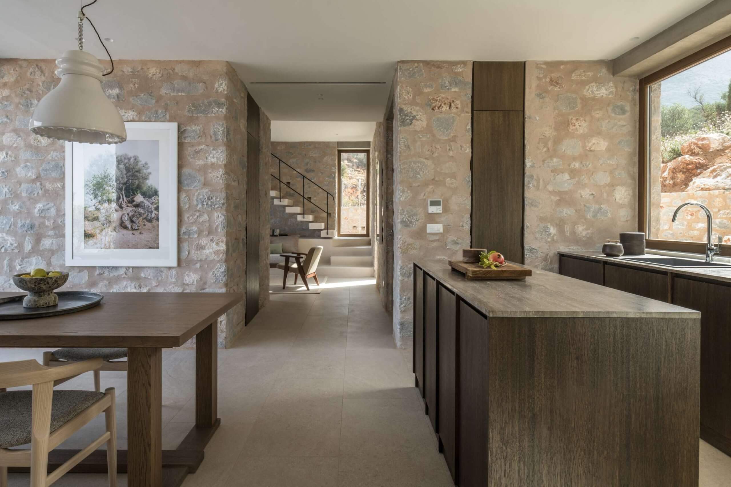 Monolithoi, Europe — Stone villa kitchen with dark timber cabinetry, natural light, travertine countertops, and textured ochre walls opening to living spaces and landscape views.