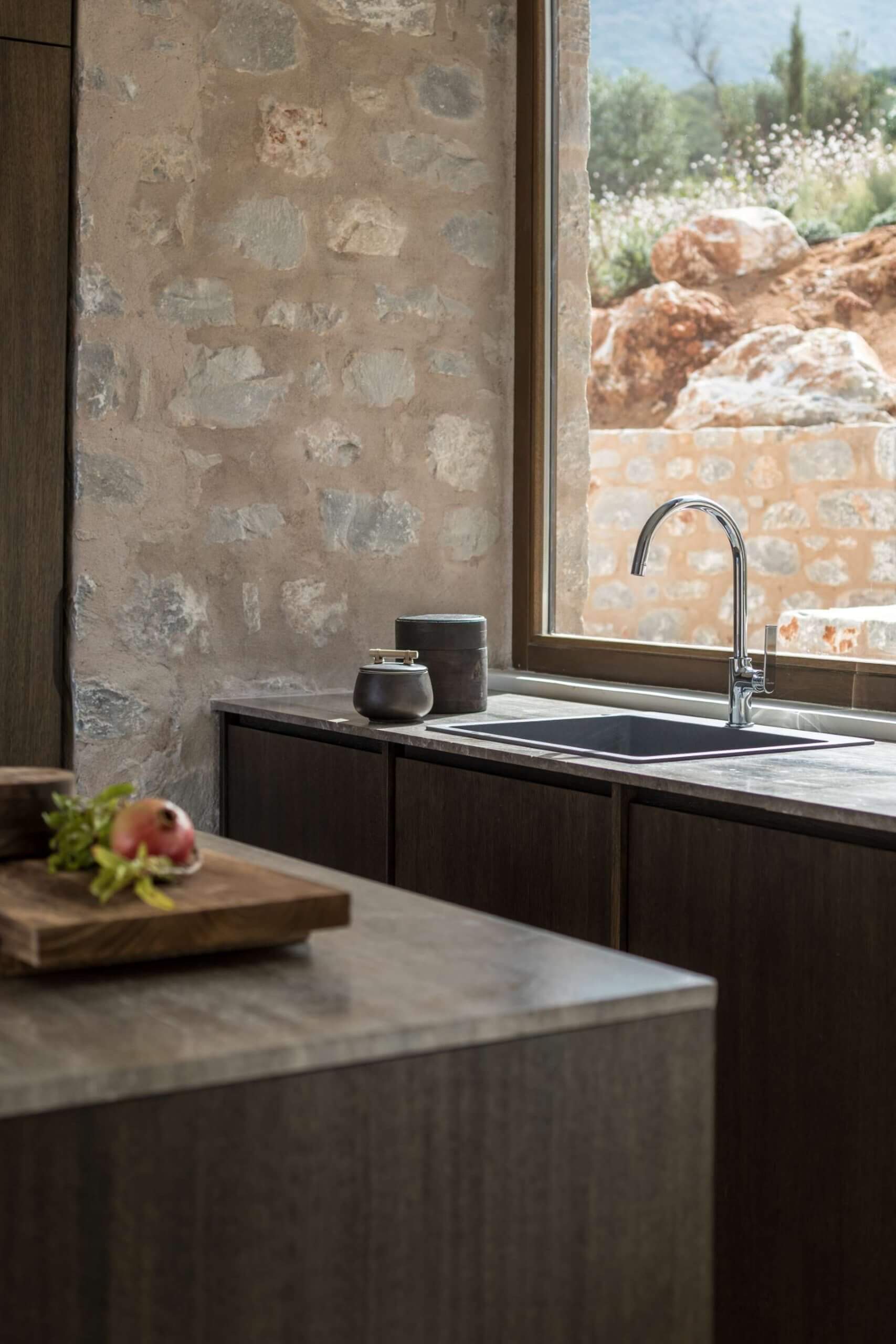 Monolithoi, Europe — Minimalist kitchen with stone sink, dark cabinetry, weathered plaster wall, and panoramic view of rocky landscape and Mediterranean garden.
