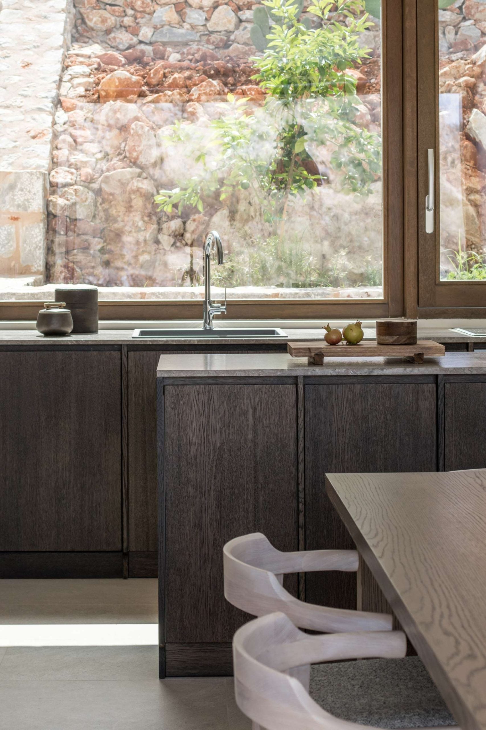 Monolithoi, Europe — Dark wood kitchen cabinetry with stone countertop, chrome faucet, and expansive window overlooking rustic stone exterior wall and climbing vines.
