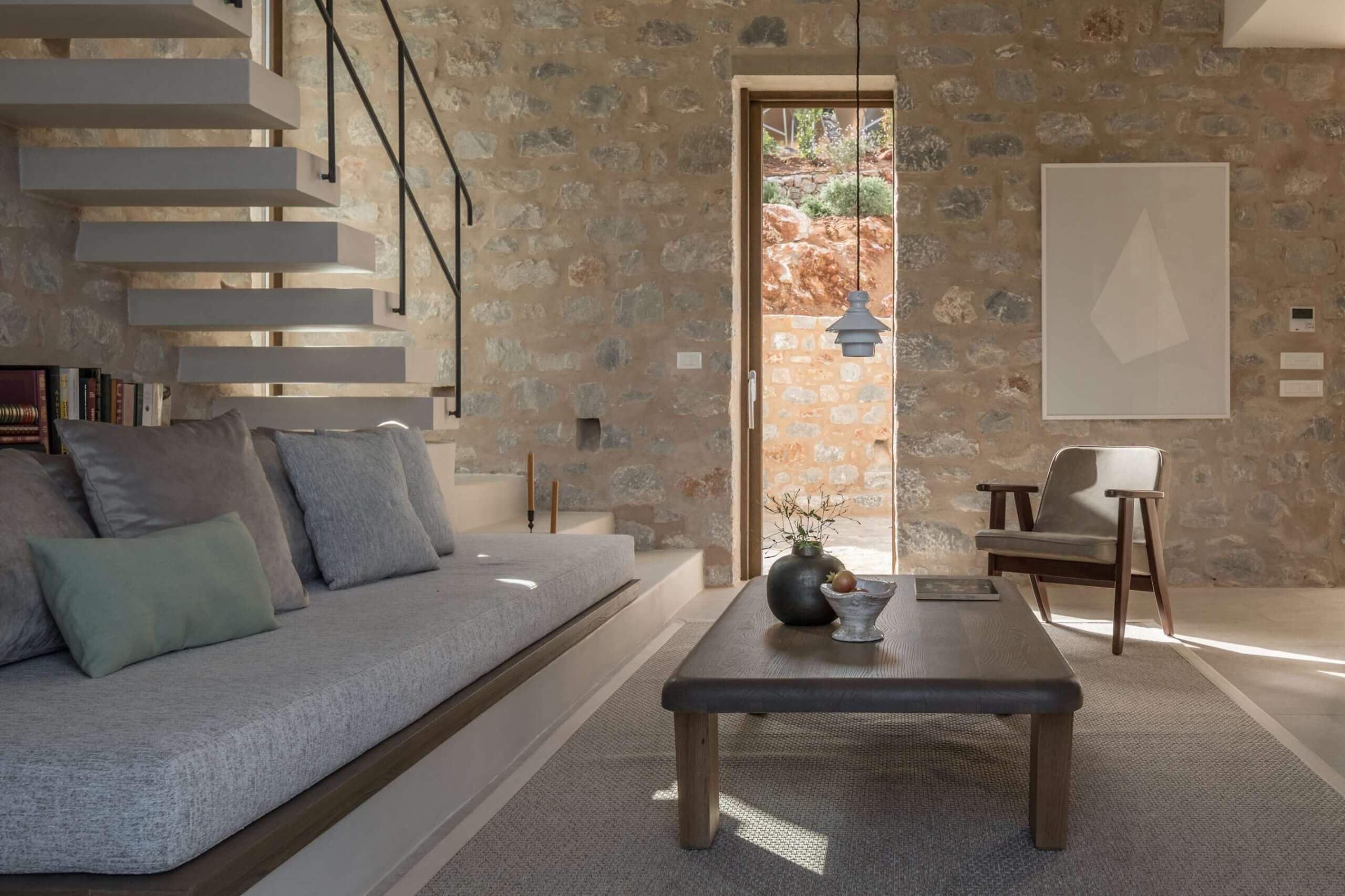 Monolithoi, Europe — Stone-walled living room with floating staircase, linen sofa, wooden furniture, and terracotta courtyard view through glass doors.