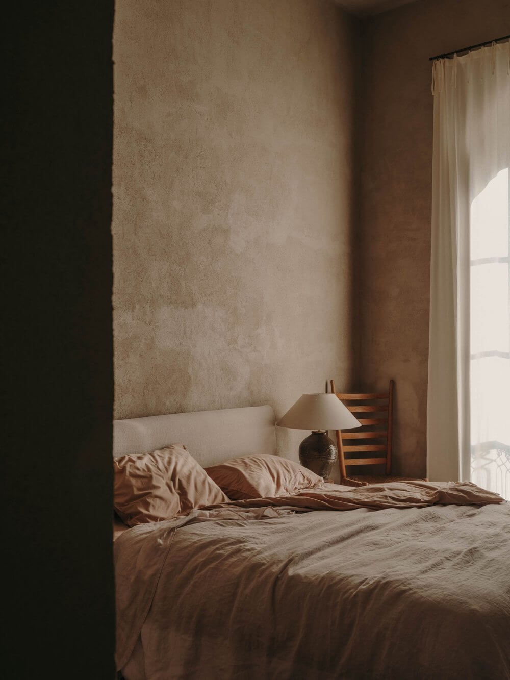 Casa Soleto, Europe — Minimalist bedroom with textured terracotta walls, cream linen bedding, wooden chair, ceramic lamp, and natural light from tall windows with sheer curtains.