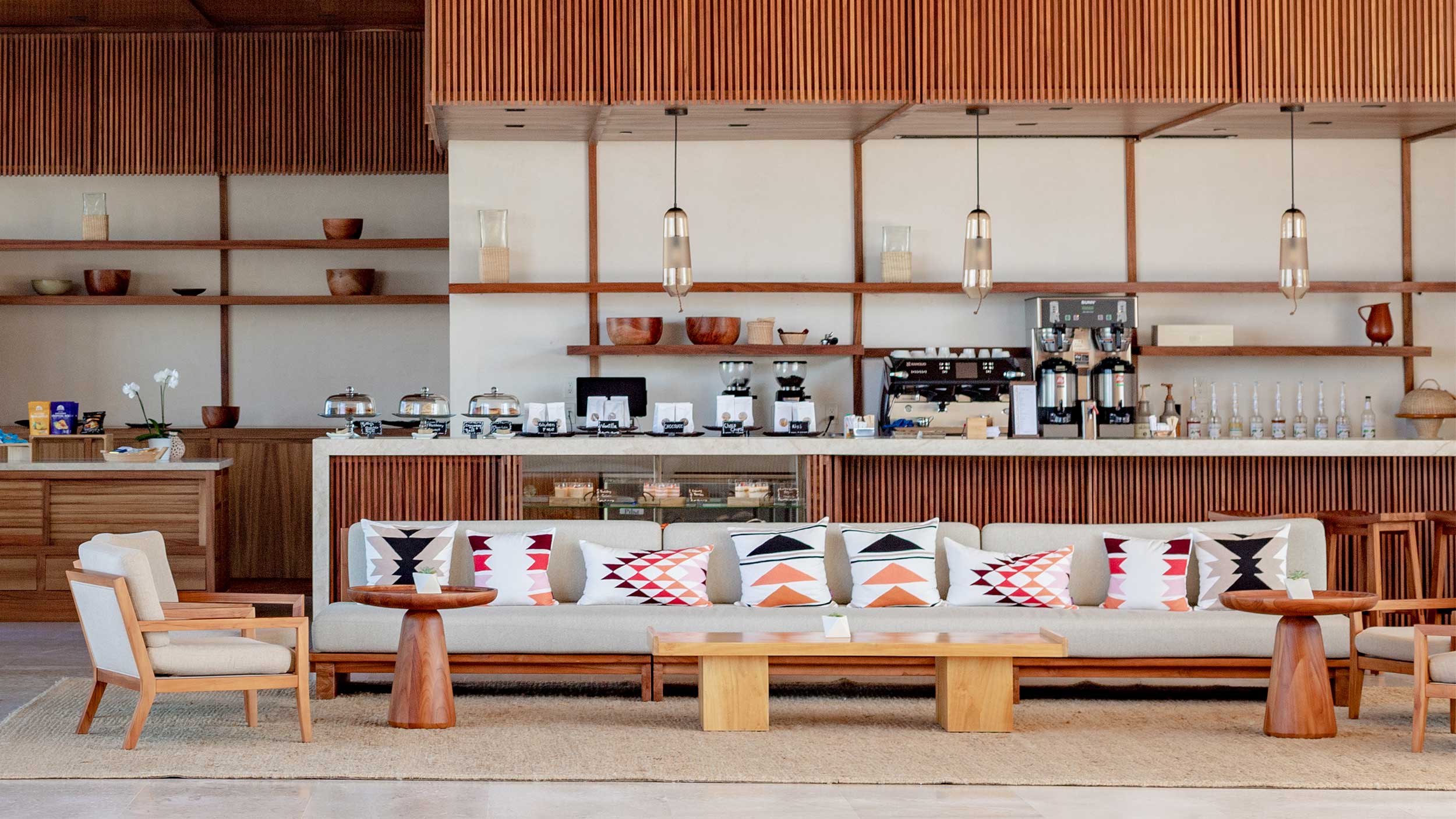 Nobu Cafe, Mexico — Warm wood shelving and pendant lights above a cream linen sofa with geometric pillows, espresso bar, mid-century modern seating, and terracotta ceramic accents.