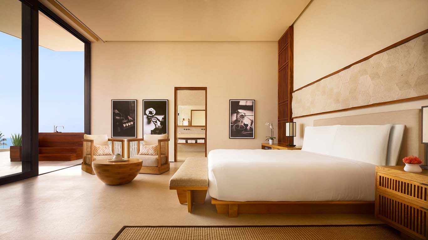 Nobu Los Cabos, Mexico — Minimalist oceanview suite with natural wood furnishings, white bedding, black photography, and floor-to-ceiling glass doors opening to terrace.