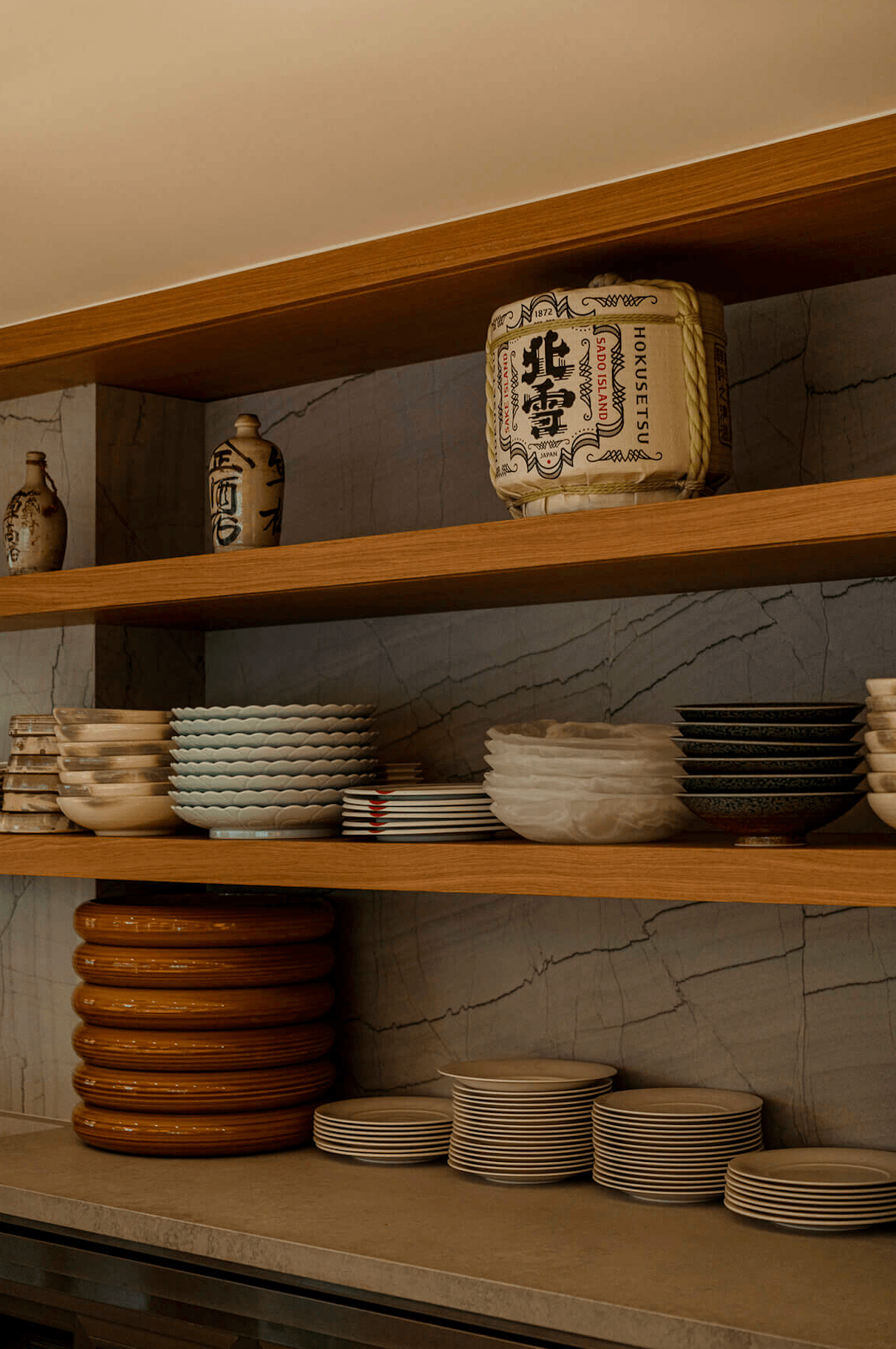 Nobu Warsaw, Poland — Open wooden shelving with stacked ceramics, wooden plates, and Japanese sake bottles against marble backsplash.