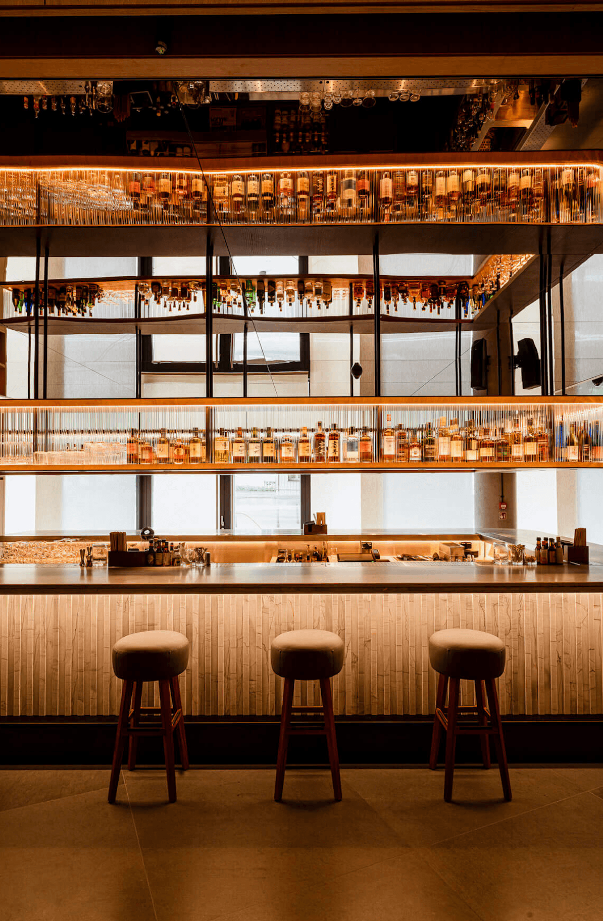 Nobu Warsaw, Poland — Multi-level bar with wood-paneled counter, brass railings displaying bottles, three leather stools, and floor-to-ceiling windows overlooking cityscape.