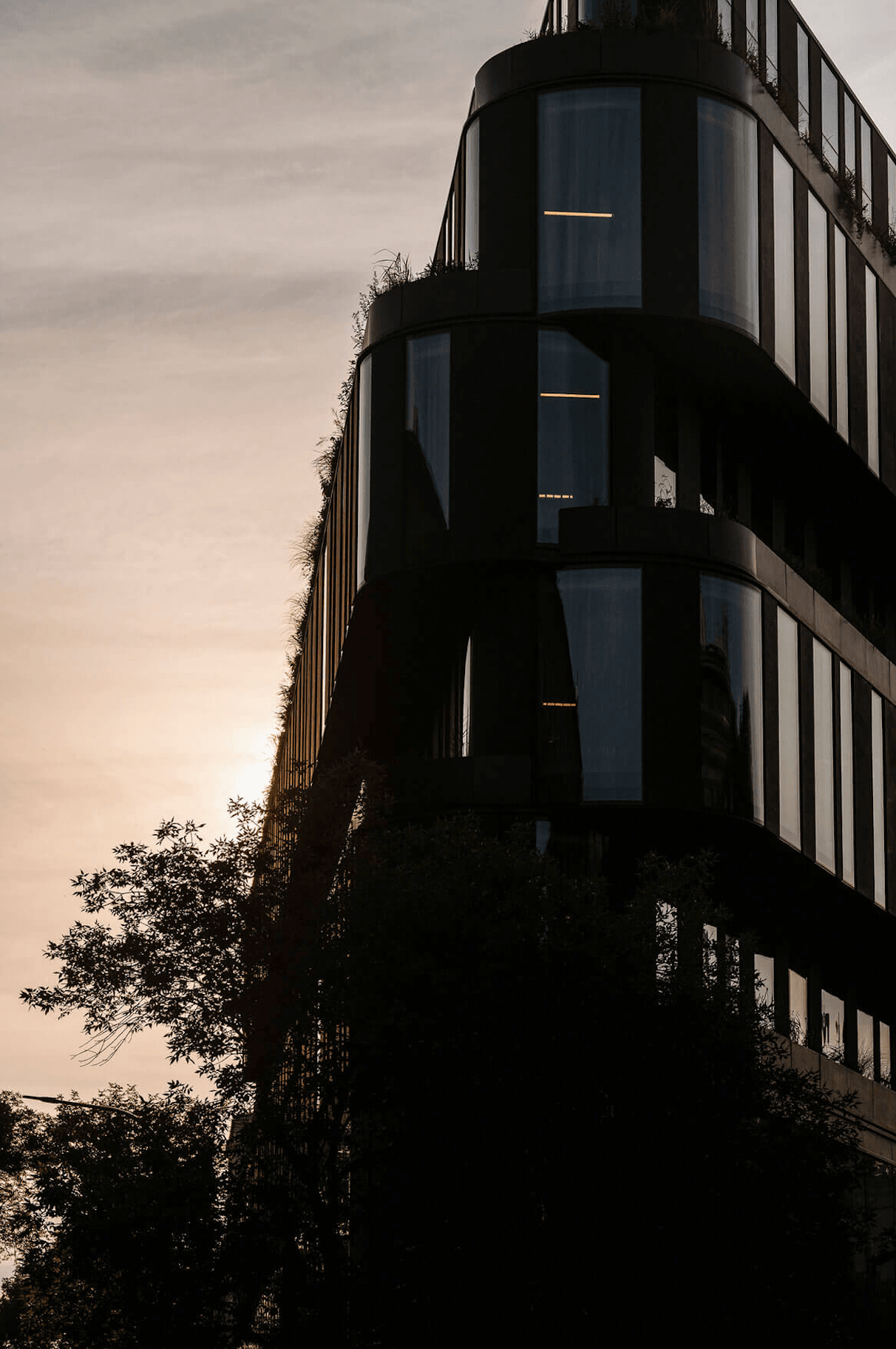 Nobu Warsaw, Poland — Curved glass facade with warm interior lighting at dusk, modernist cylindrical tower framed by surrounding foliage and overcast sky.
