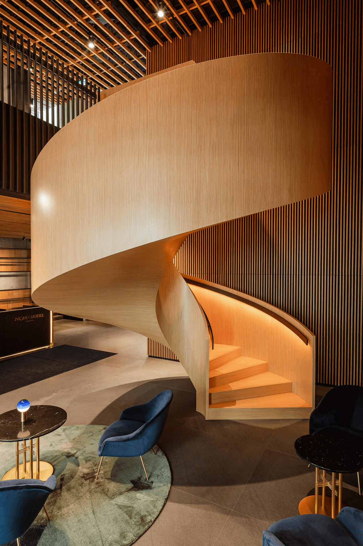Nobu Warsaw, Poland — Sculptural wooden spiral staircase with curved walls, warm timber finishes, and vertical slat detailing in contemporary luxury lobby.
