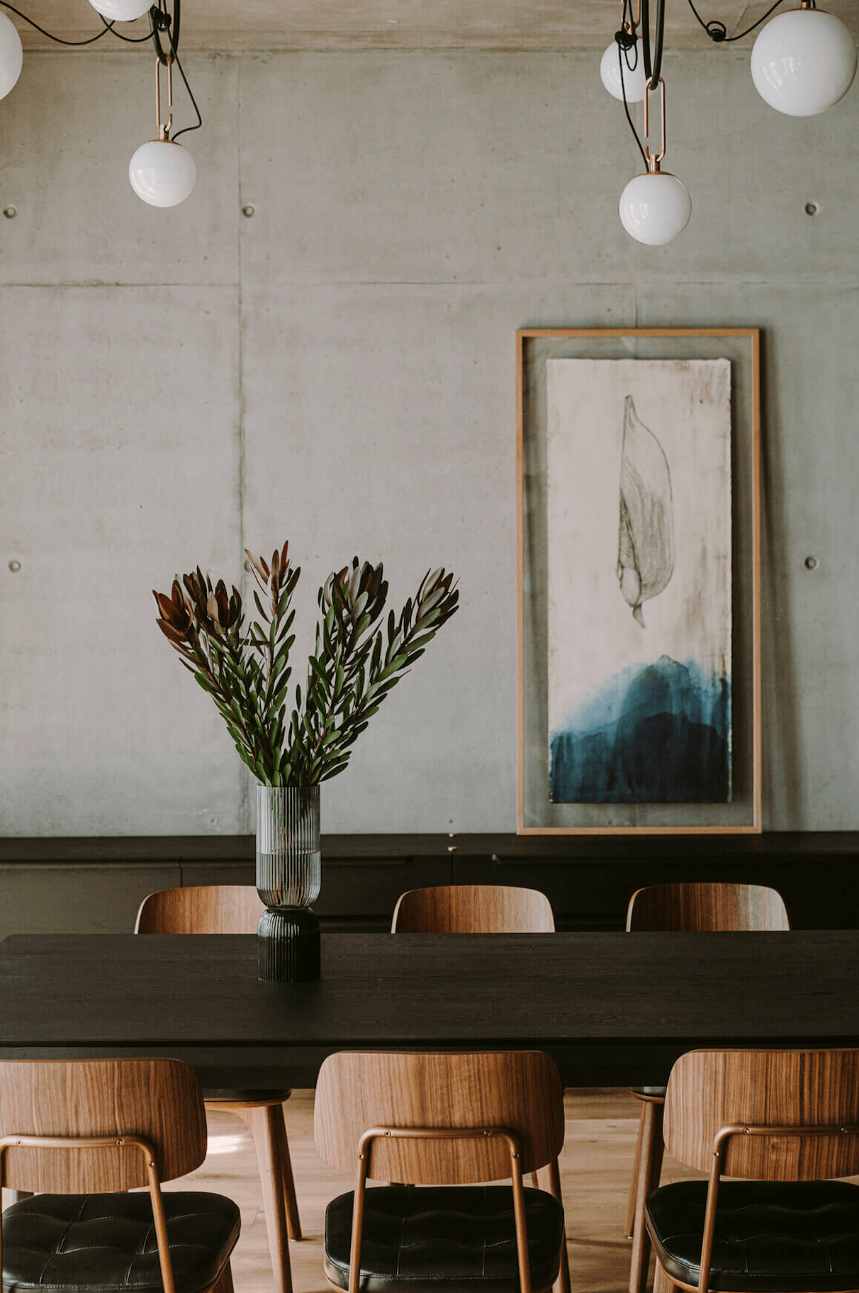Nobu Warsaw, Poland — Minimalist dining room with concrete walls, wooden chairs, pendant globe lights, and botanical artwork above dark table.