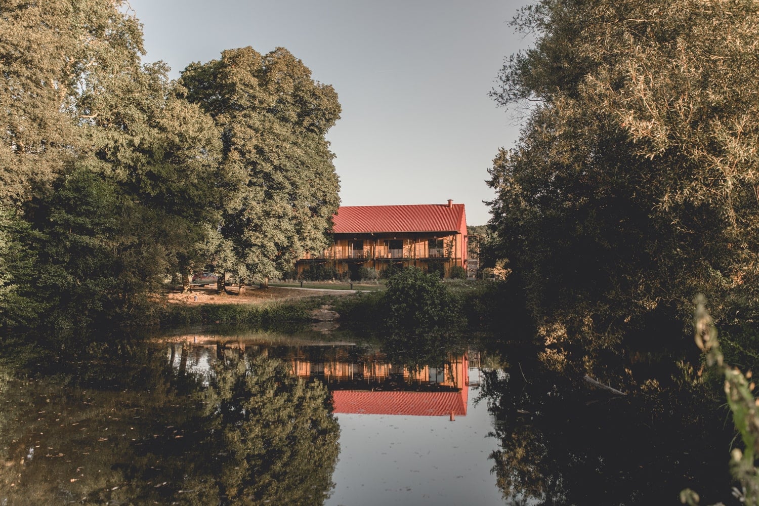Le Barn, Europe — Wooden lodge with red metal roof reflected in still water, framed by golden autumn trees at dusk.
