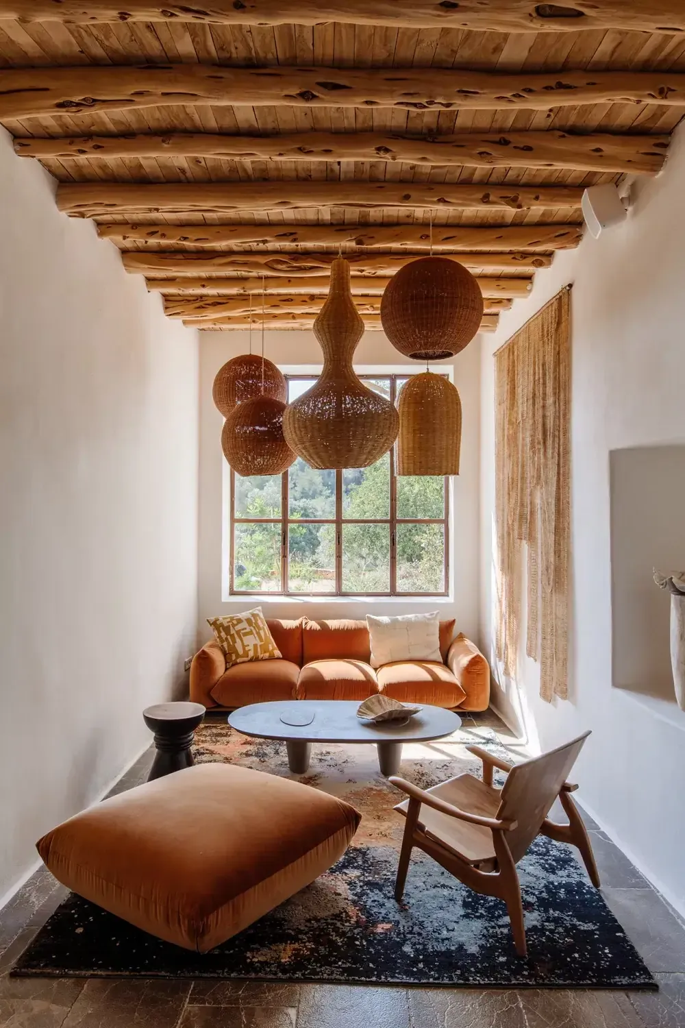 Finca La Inika, Balearic Islands — Woven pendant lights hang beneath wooden beam ceiling above rust-orange sofa and natural linen seating by large gridded window with rural views.