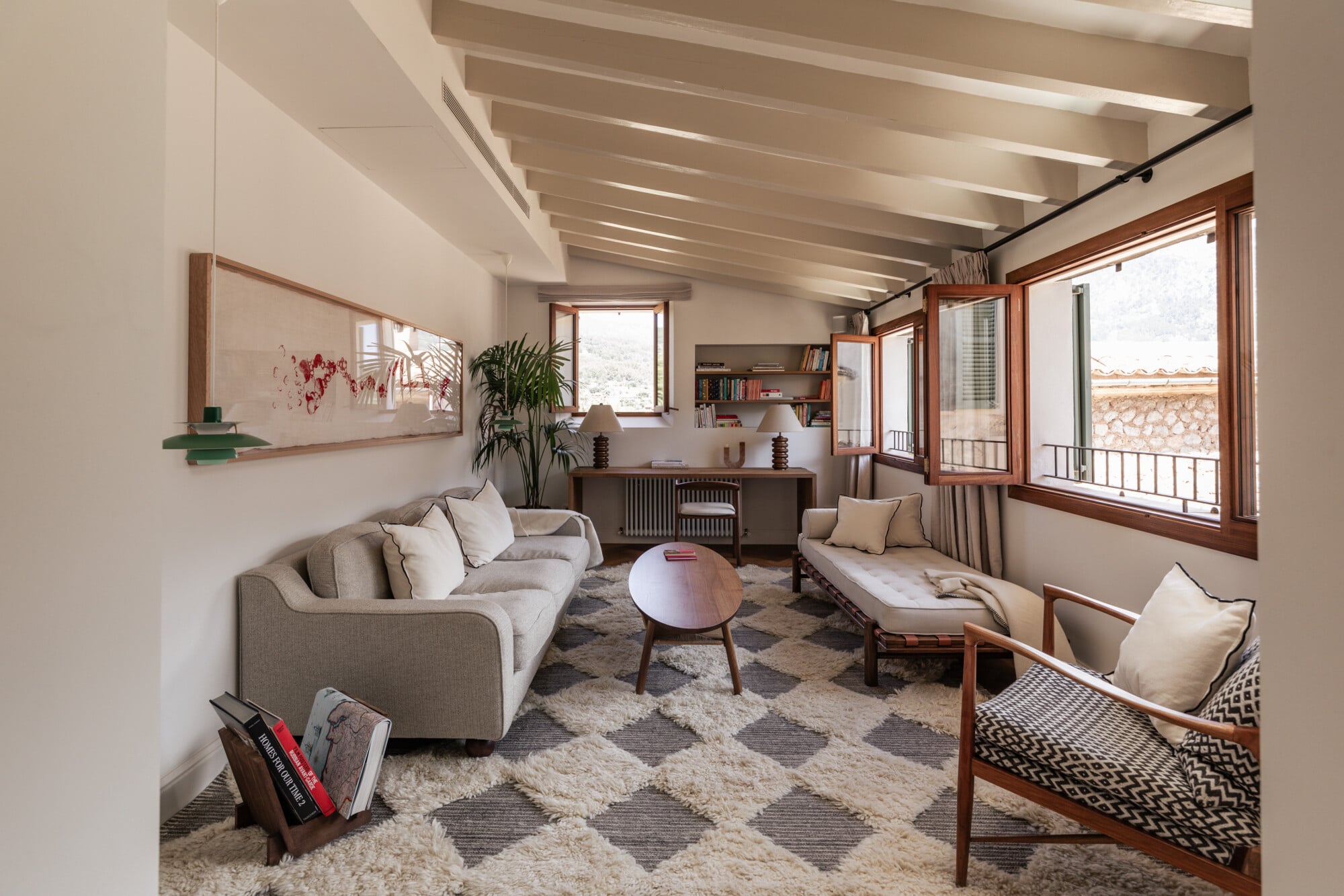 Mon Cor, Balearic Islands, Spain — Modern living room with exposed beam ceiling, warm wood-framed windows, neutral furnishings, geometric patterned rug, and Mediterranean landscape views.
