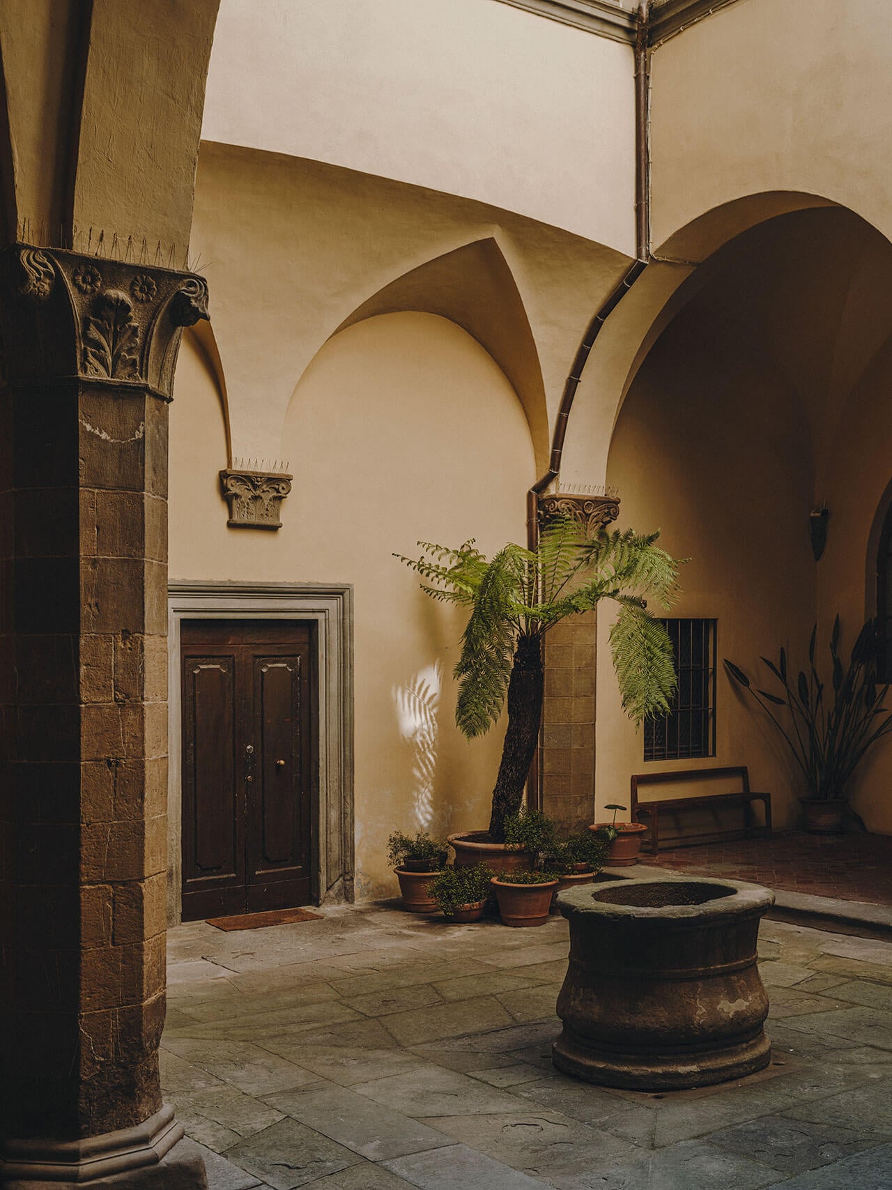 Numeroventi, Europe — Ochre courtyard with Corinthian columns, arched loggia, terracotta planters, central well, and warm Mediterranean light casting shadows on stone floors.