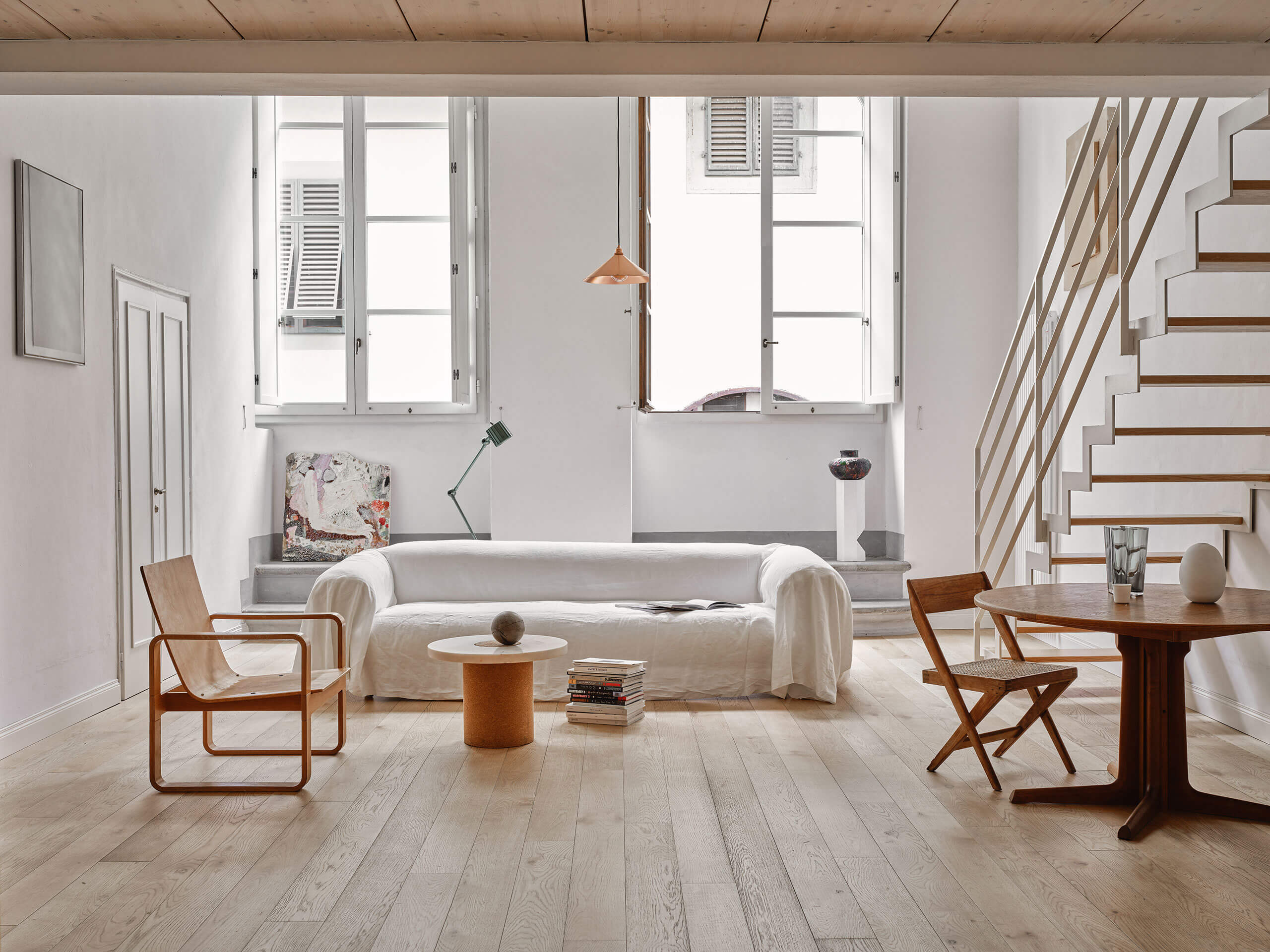 Numeroventi, Europe — Minimalist loft with white sofa, wooden mid-century chairs, pale oak flooring, steel-frame windows, and floating staircase in natural light.