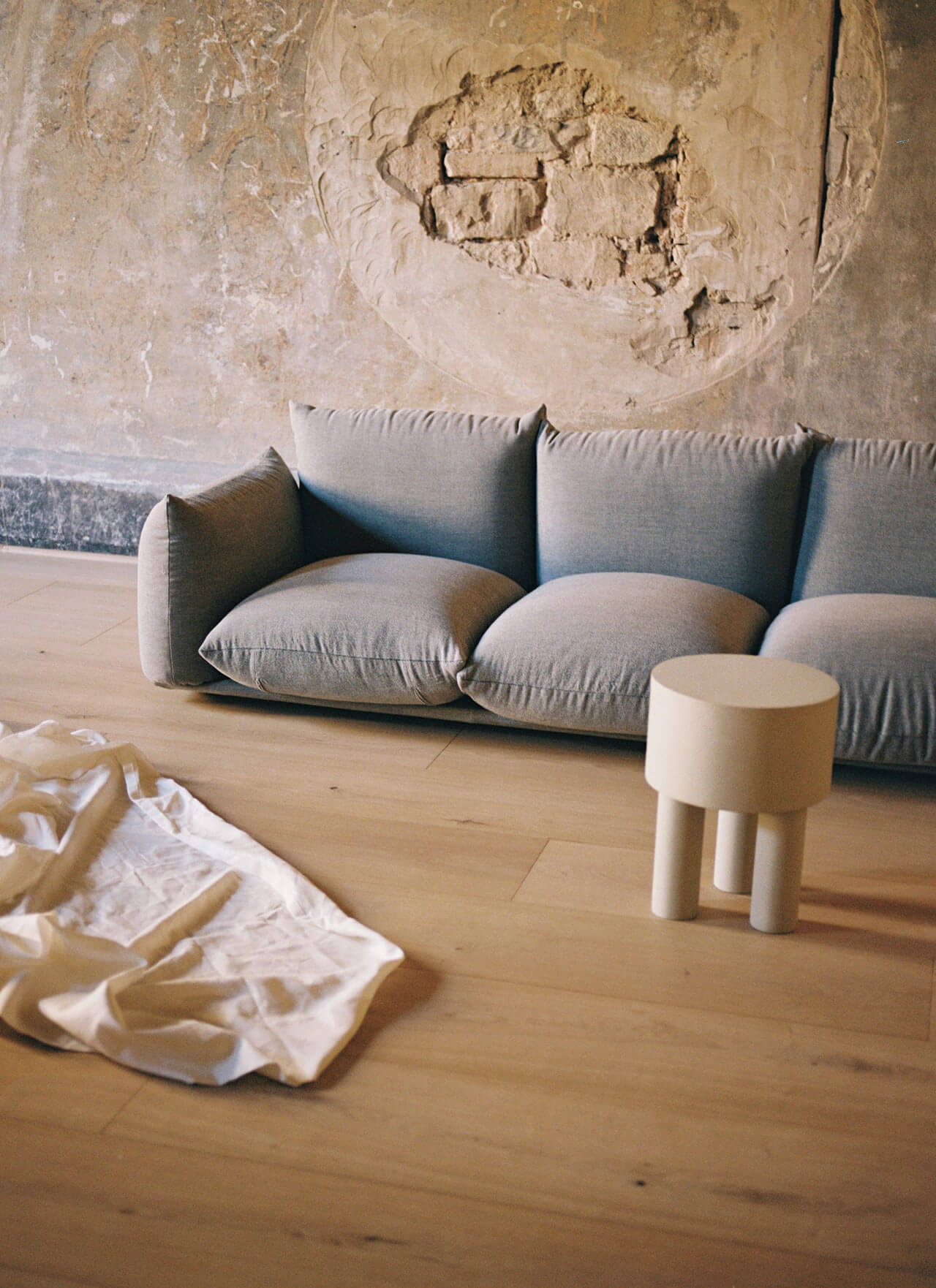 Numeroventi, Europe — Minimalist living room with pillowed gray sofa, wooden stool, natural light, and textured plaster wall with sculptural relief detail.