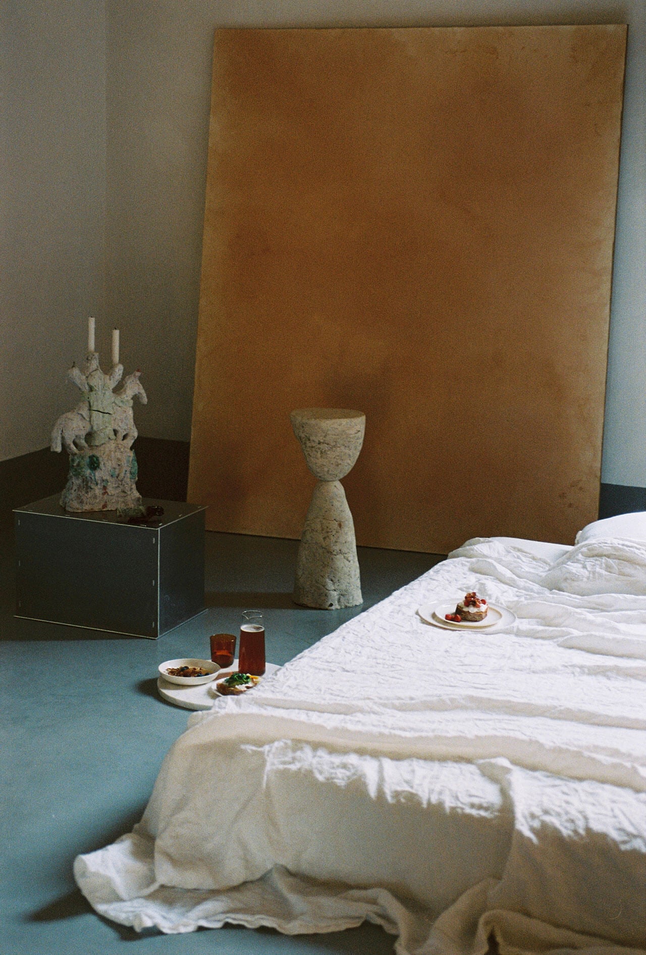 Numeroventi, Europe — Minimalist bedroom with white linens, ochre canvas backdrop, stone sculptural vessels, and seafoam floor creating serene luxury interior.