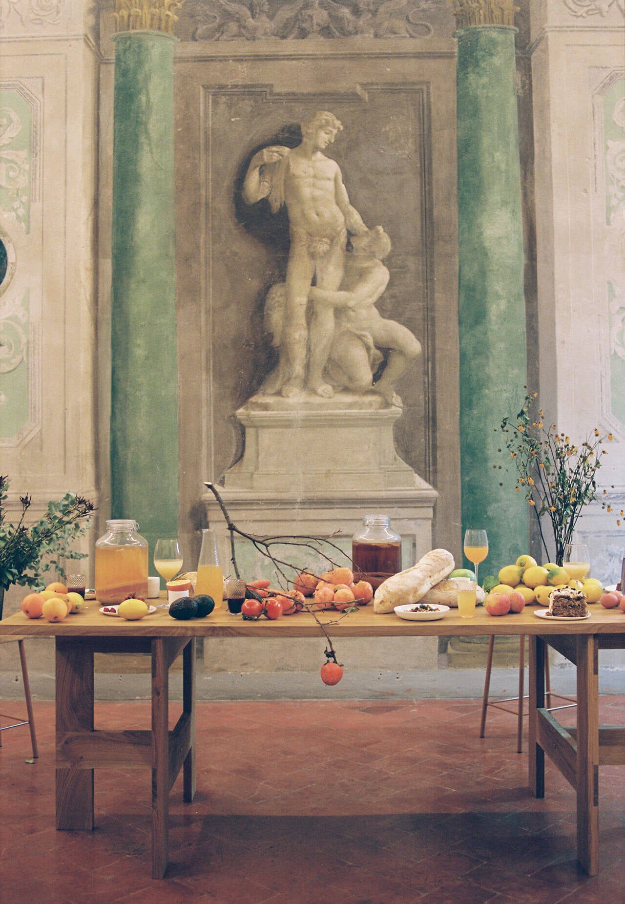 Numeroventi, Europe — Neoclassical statue niche with green marble columns, wood table styled with seasonal fruits, bread, and drinks in classical interior gallery space.