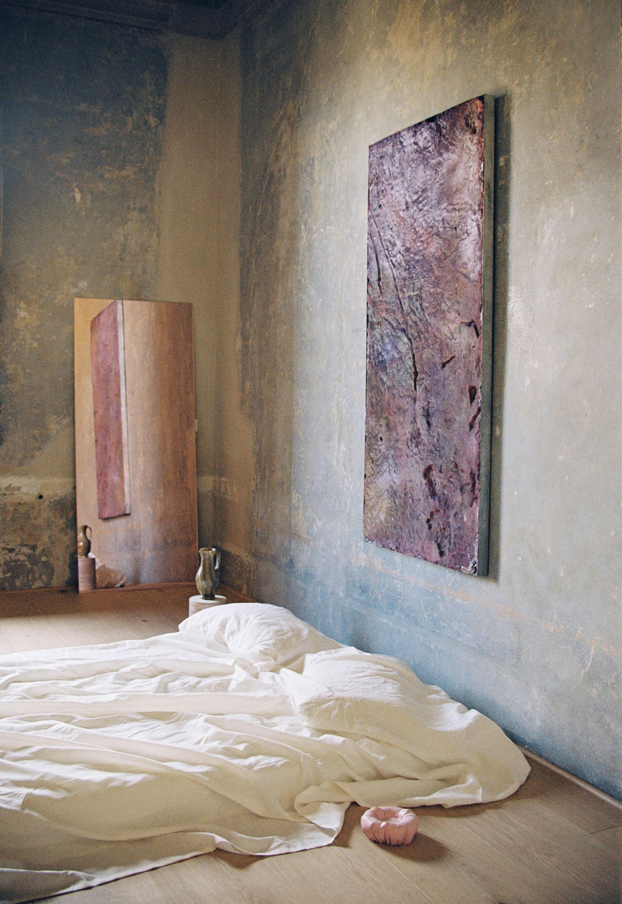 Numeroventi, Europe — Minimalist bedroom with cream linens, weathered plaster walls, and abstract textile art in muted tones.