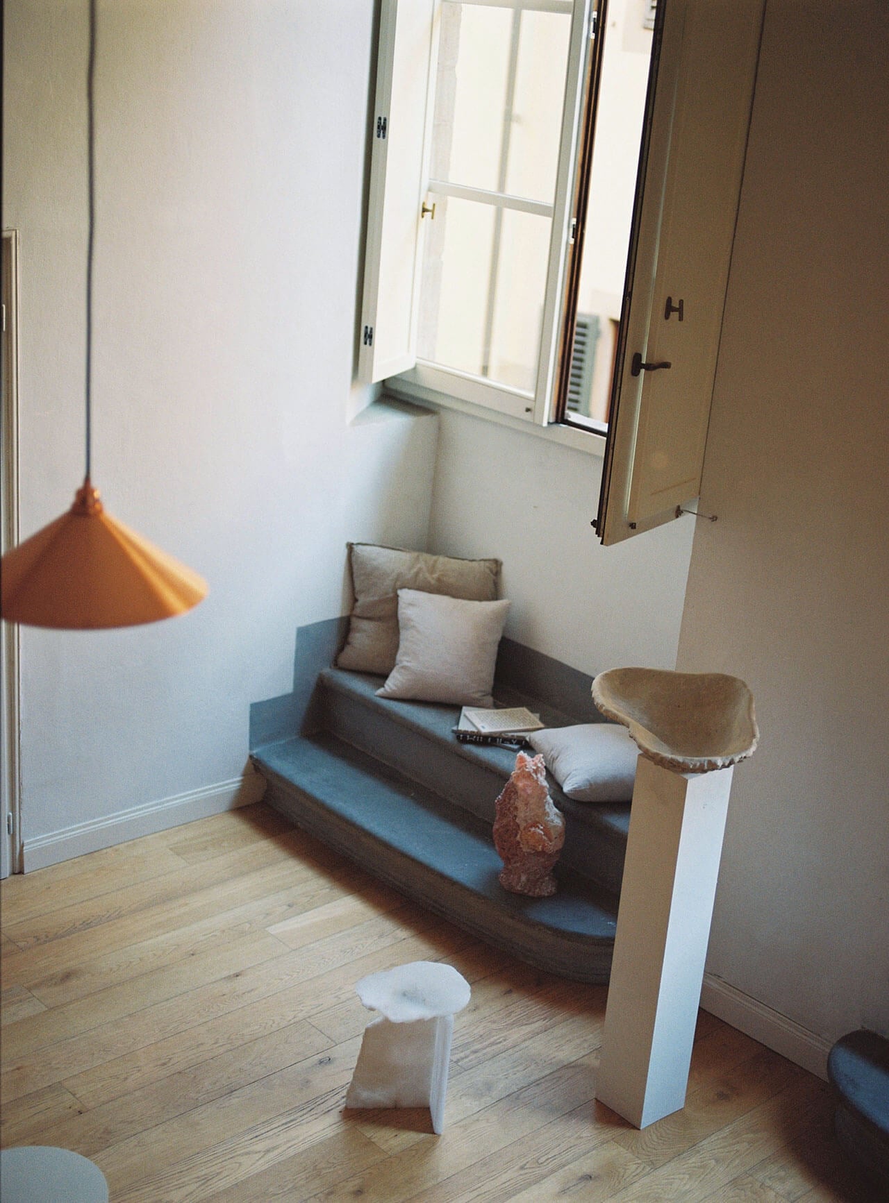 Numeroventi, Europe — Minimalist window nook with gray upholstered bench, linen pillows, pendant lamp, wooden floors, and natural light from casement window.