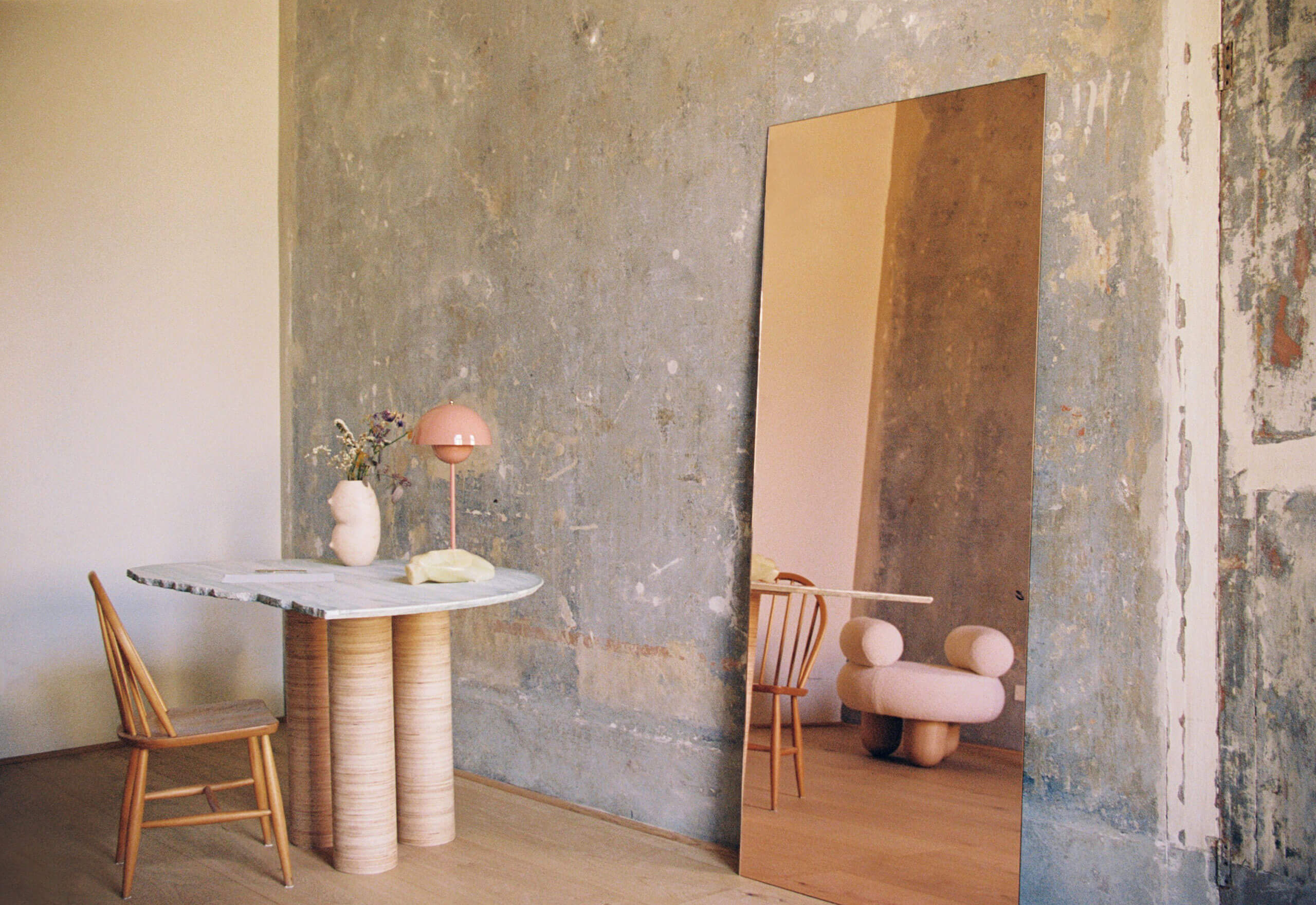 Numeroventi, Europe — Concrete gallery wall with brass mirror, wooden pedestal table, pink sculptural chair, and minimalist tabletop styling.