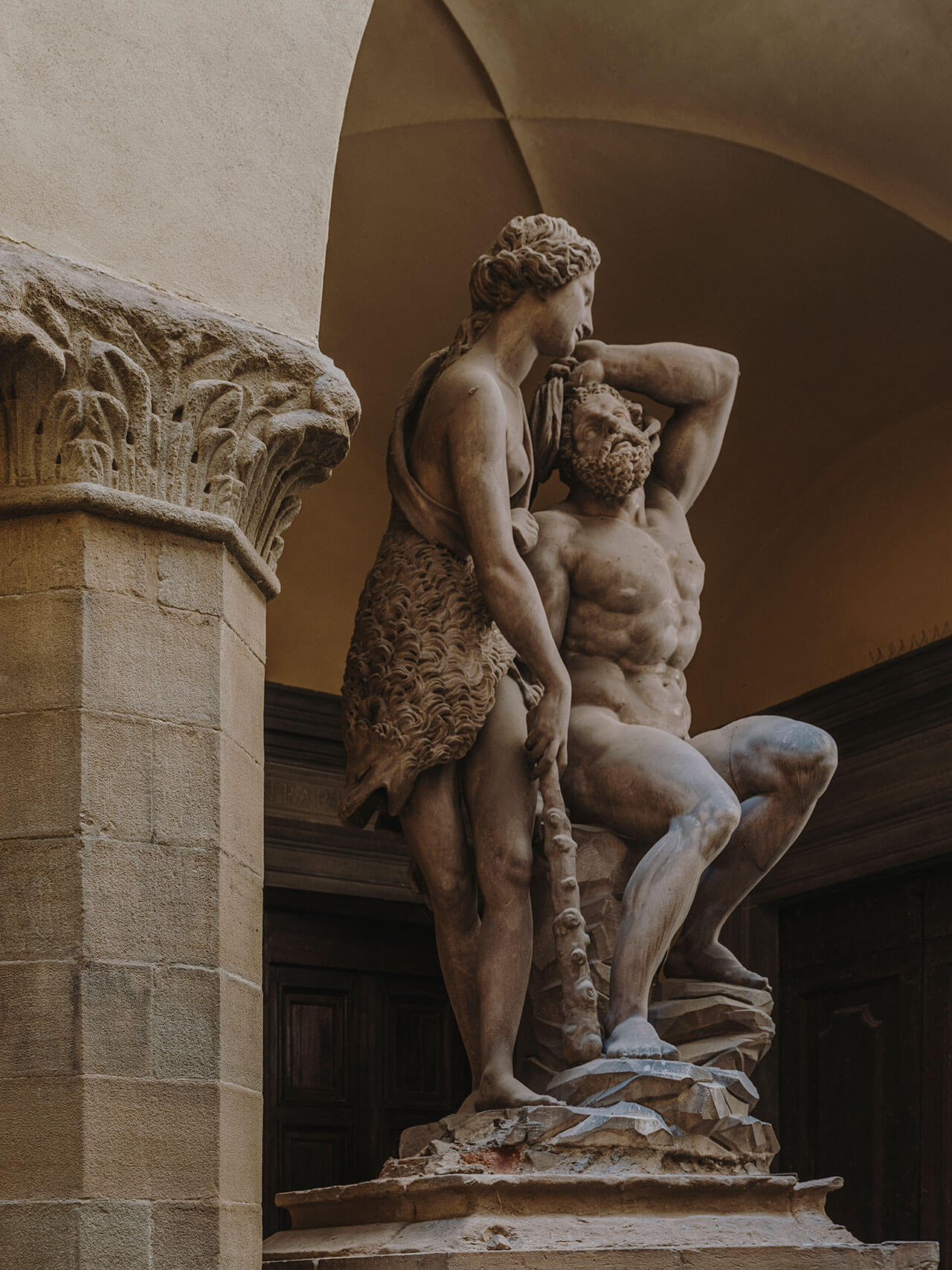 Numeroventi, Europe — Classical stone sculpture of intertwined figures in arched courtyard with terracotta walls and ornamental capital detail.