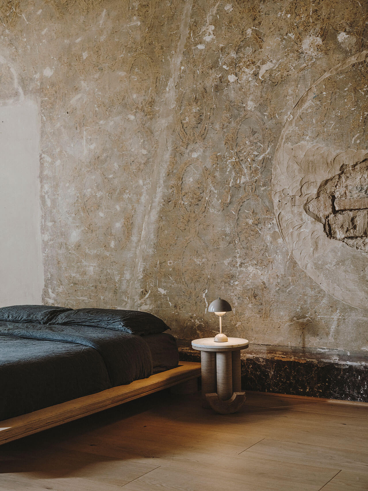 Numeroventi, Europe — Minimalist bedroom with weathered plaster wall, low platform bed, wooden floor, and sculptural bedside lamp creating stark architectural atmosphere.