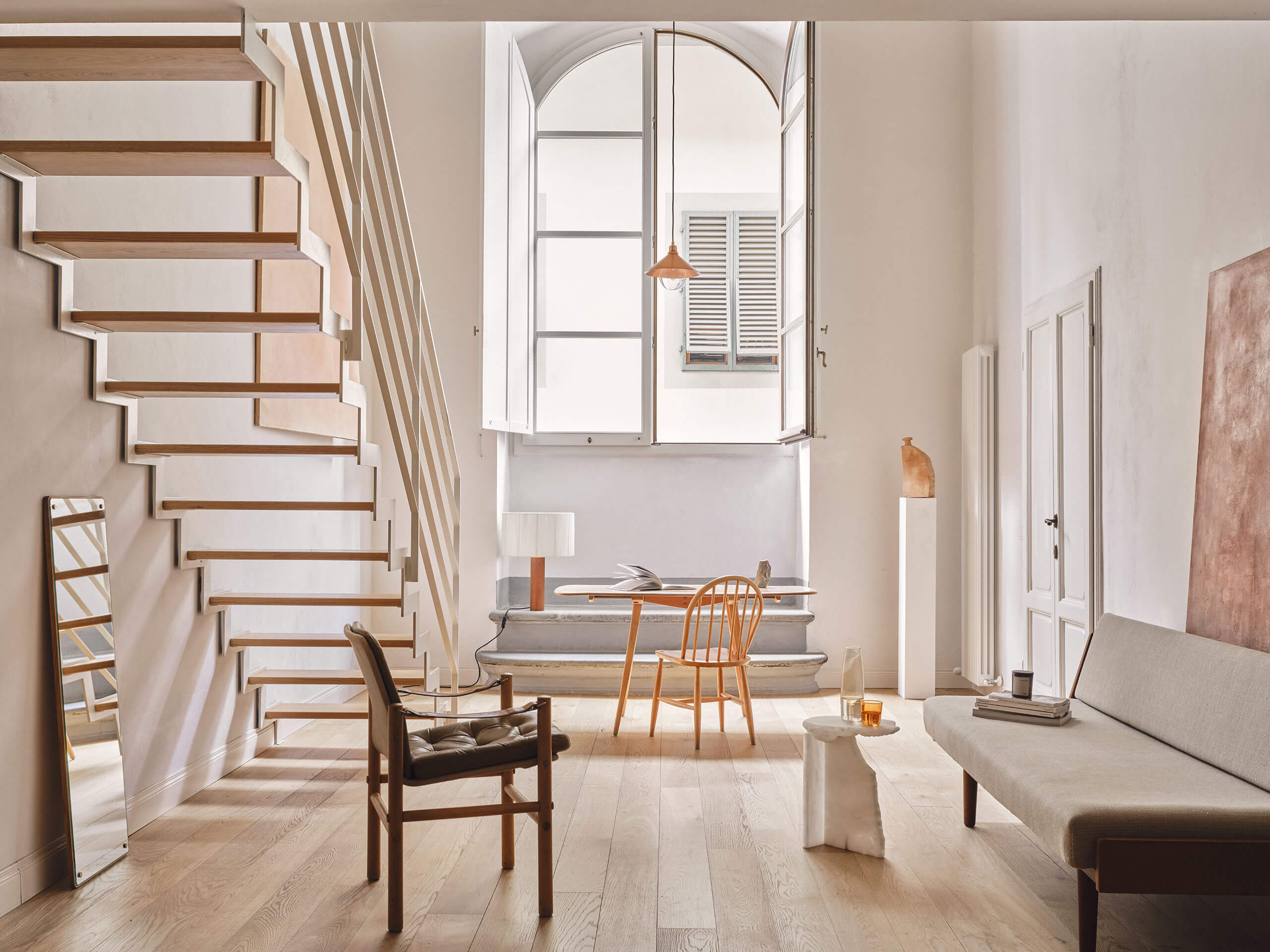 Numeroventi, Europe — Sunlit loft with floating timber stairs, arched windows, wooden furniture, and minimal furnishings in neutral palette.