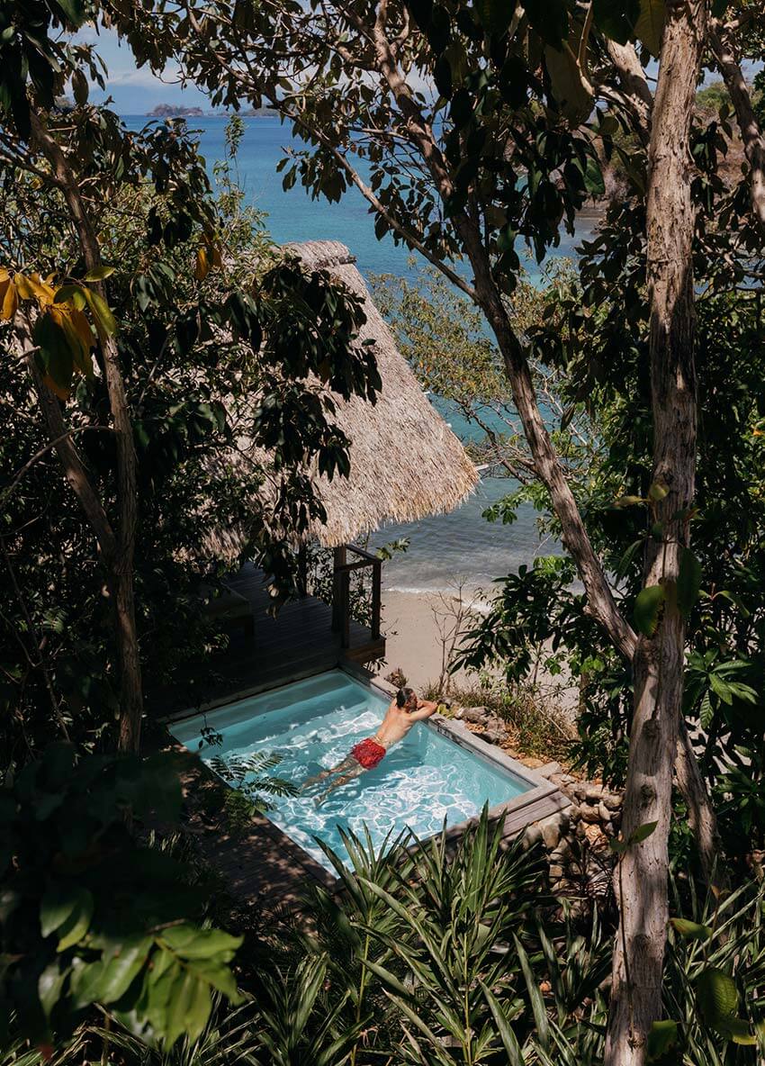Islas Secas, Panama — Person floating in turquoise pool nestled between tall trees, with thatched-roof bungalow and Caribbean Sea beyond.