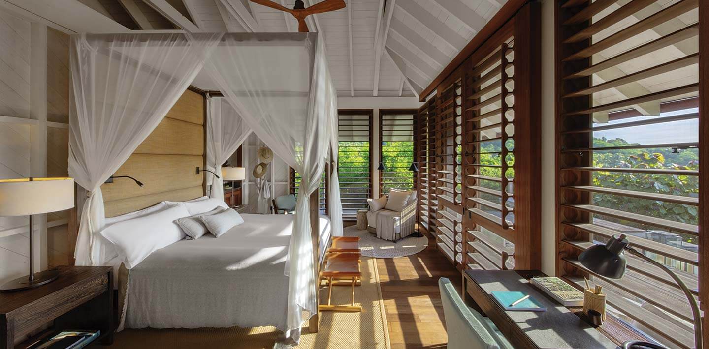 Islas Secas, Panama — Canopy bed with sheer white curtains in modern villa bedroom with wooden louver shutters, polished concrete floors, and panoramic views of tropical landscape.