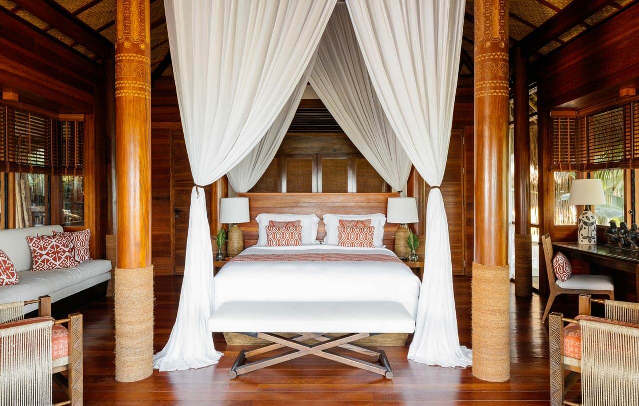 NIHI Sumba, Asia — Teak villa bedroom with four-poster canopy bed, white drapes, carved wooden columns, coral patterned cushions, and tropical hardwood flooring.