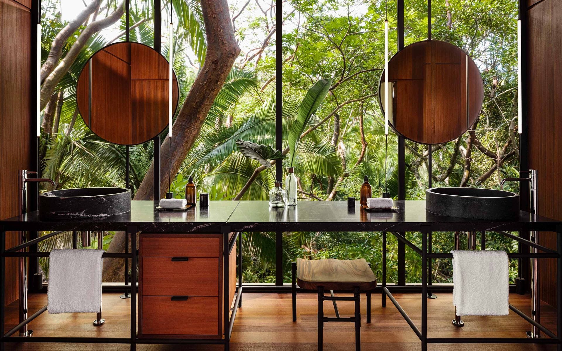 One&Only Mandarina, Mexico — Dual black stone vessel sinks with circular mirrors, dark vanity counter, tropical forest views through floor-to-ceiling windows, warm wood and modern minimalist design.