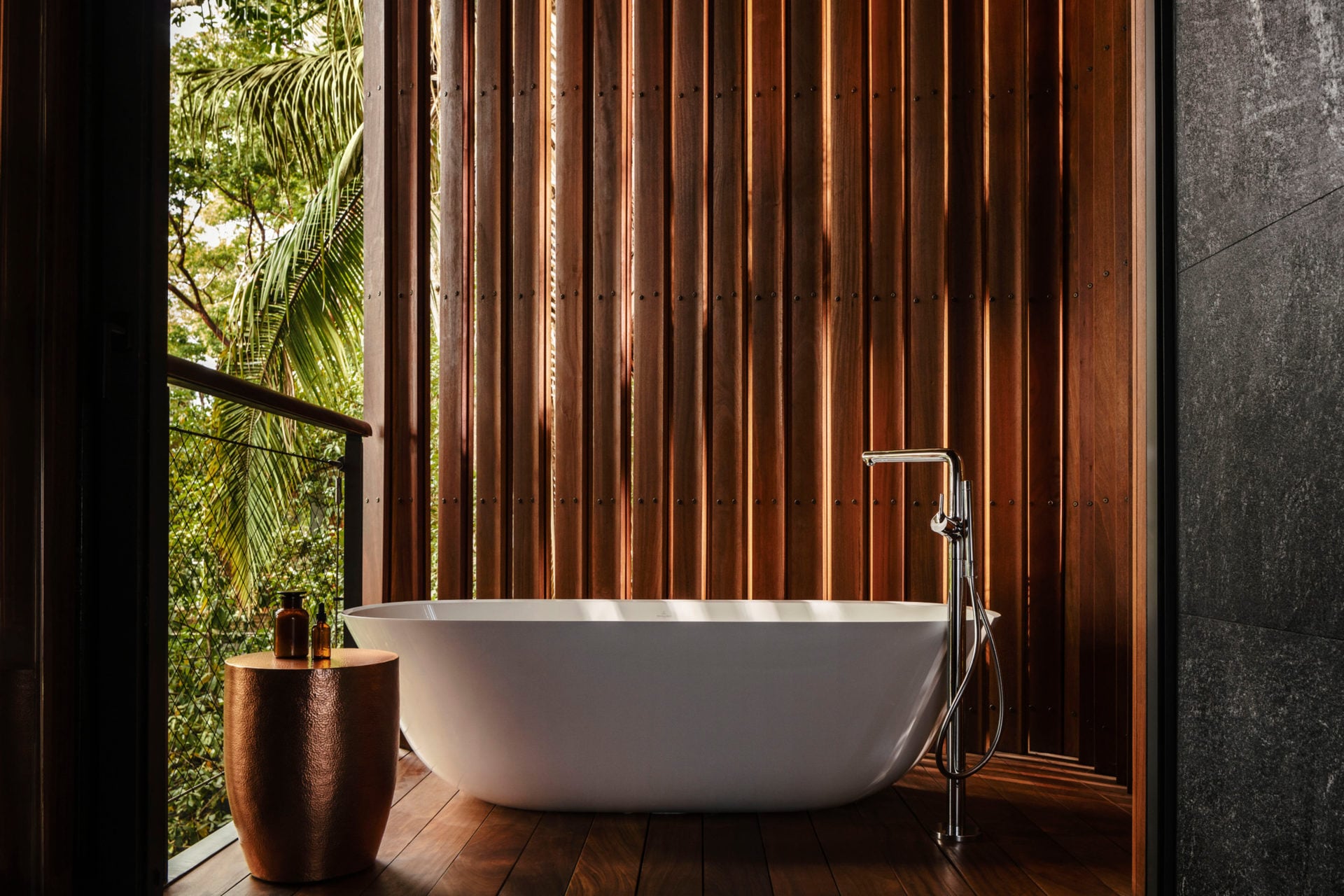 One&Only Mandarina, Mexico — Freestanding white bathtub on wooden deck with vertical timber screen, chrome faucet, tropical garden view, concrete wall, natural light.