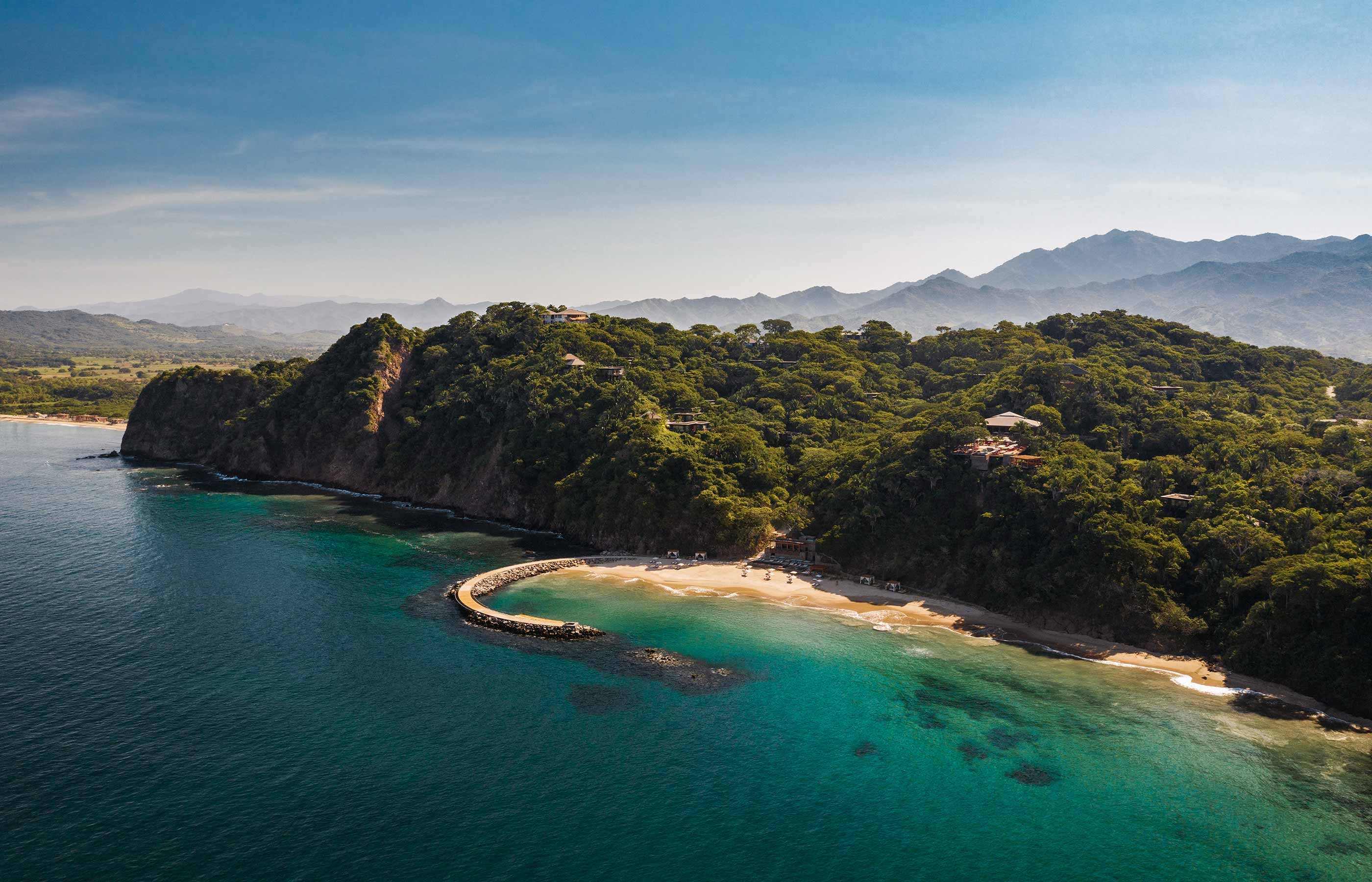 One&Only Mandarina, Mexico — Aerial view of crescent beach cove with turquoise water, dark volcanic cliffs, dense tropical forest, and mountains beyond under clear sky.
