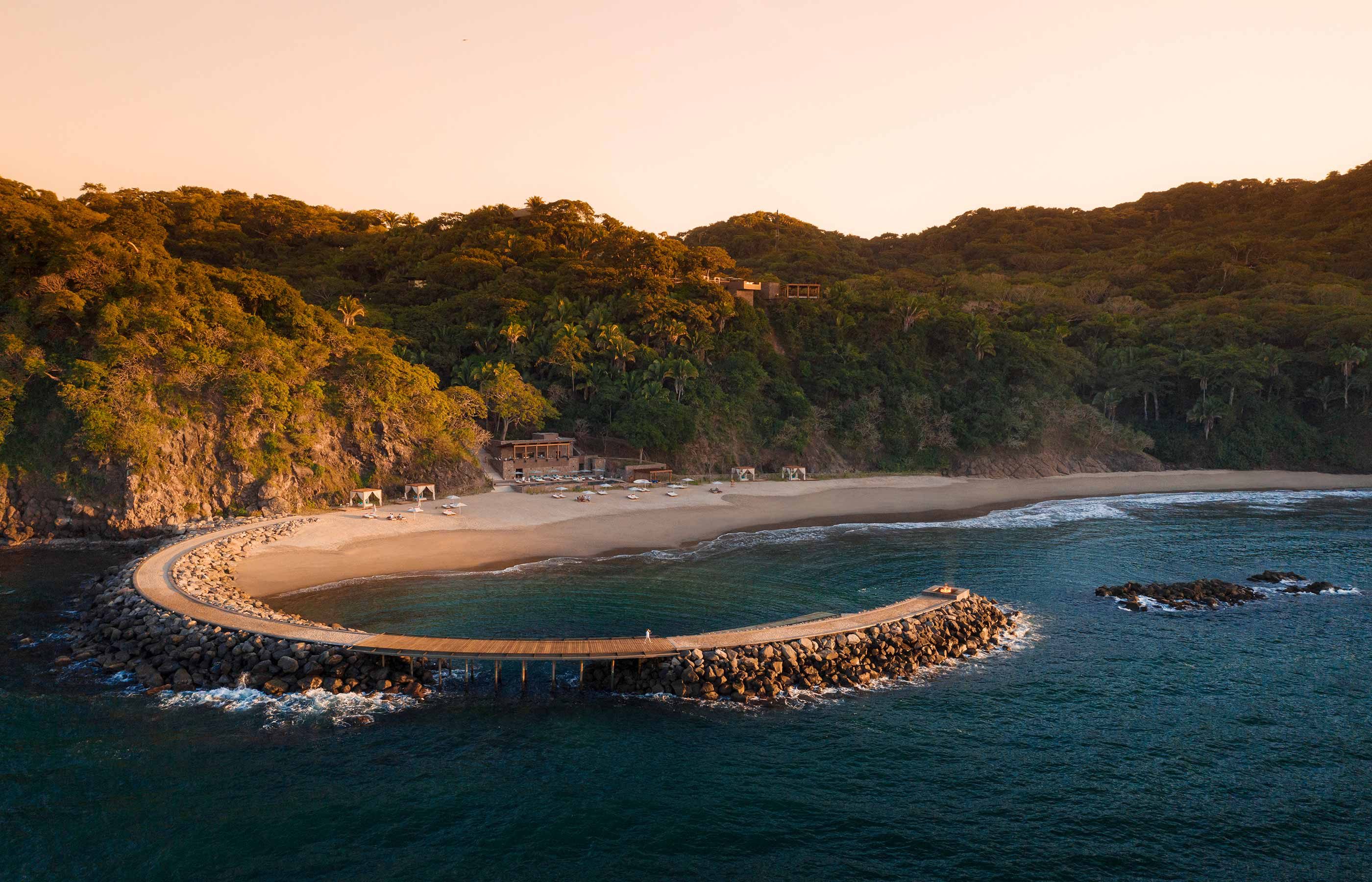 One&Only Mandarina, Mexico — curved wooden boardwalk and sandy cove with turquoise waters, jungle-covered cliffs at golden hour.