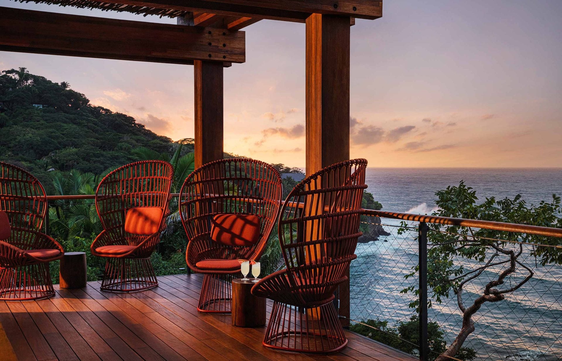 The Treetop, Mexico — Red woven chairs with orange cushions on wooden deck at sunset, overlooking ocean and forested coastline beneath timber pergola.