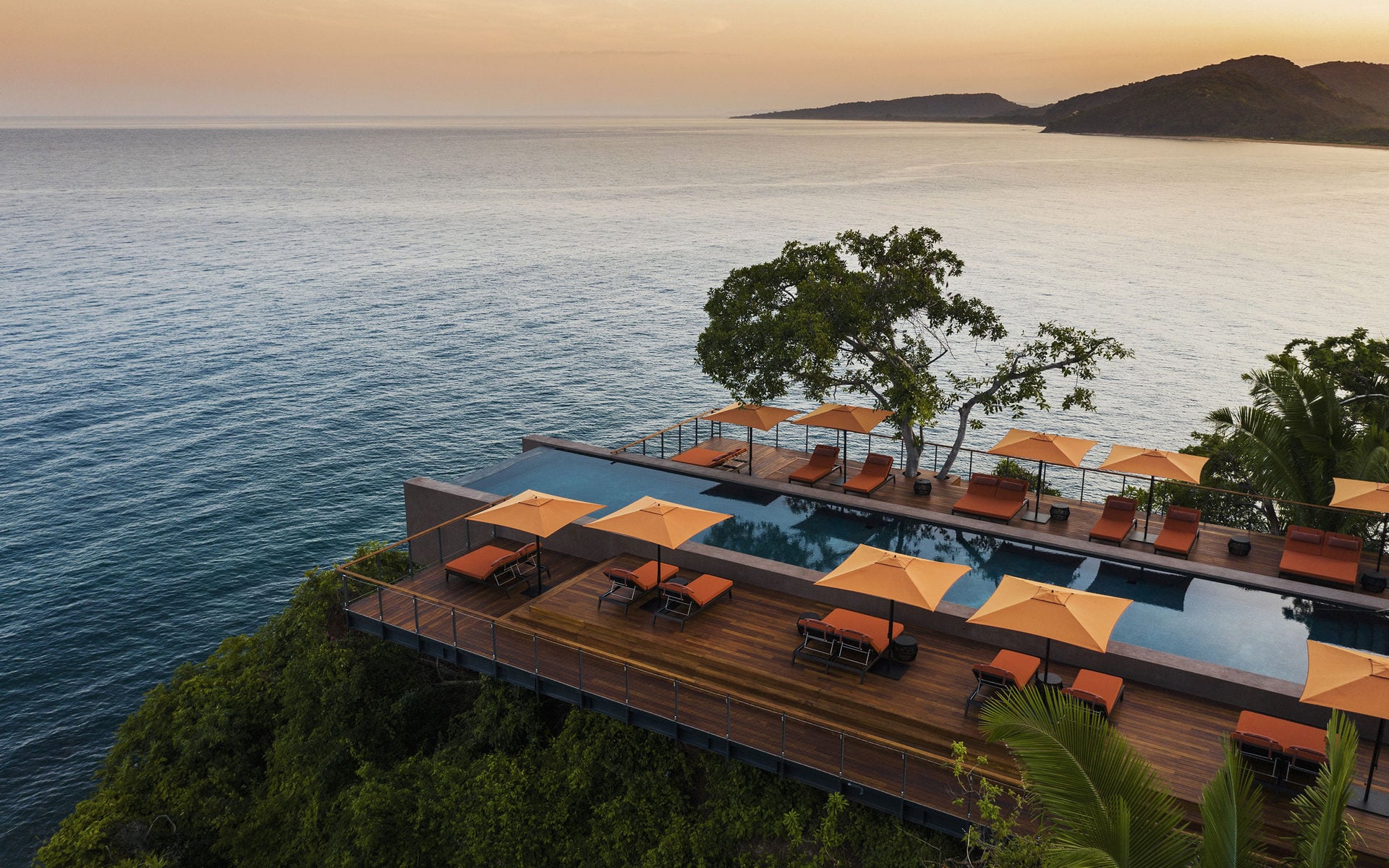 One&Only Mandarina, Mexico — Infinity pool with orange umbrellas overlooking Pacific Ocean at sunset, wooden deck, coastal cliffs, minimalist luxury resort architecture.