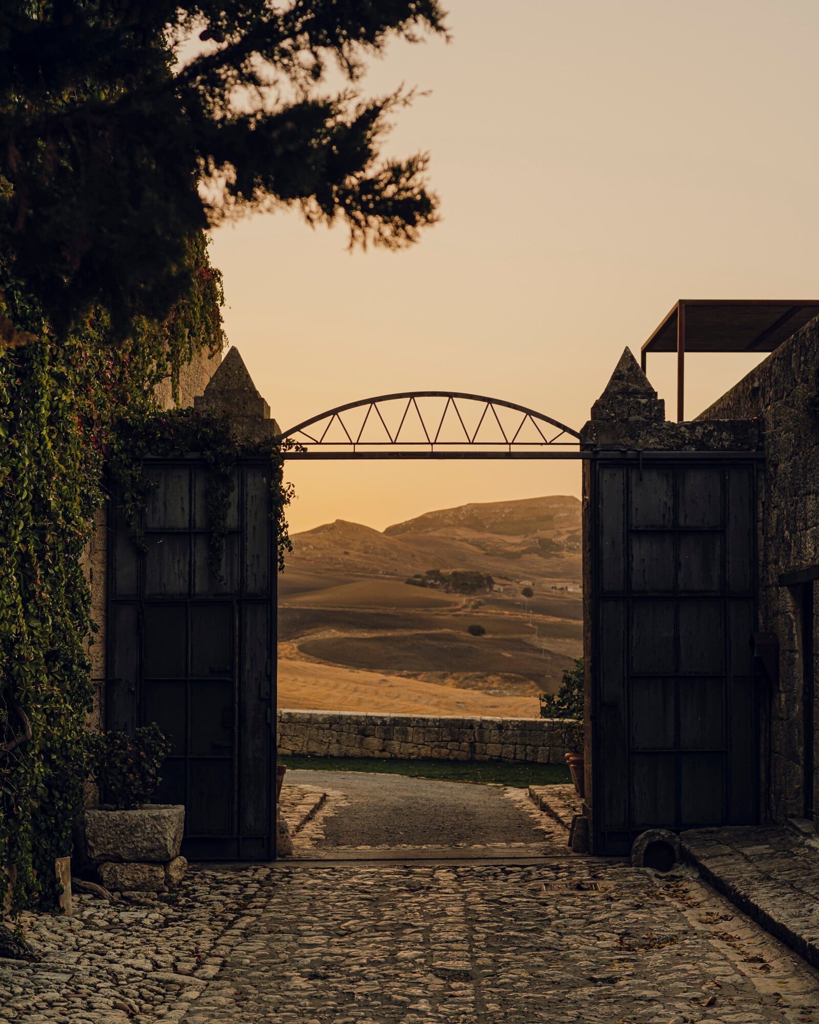 Susafa, Italy — Open ironwork gates framing golden-hour landscape, ivy-clad stone walls, arched bridge, rolling hills beyond.