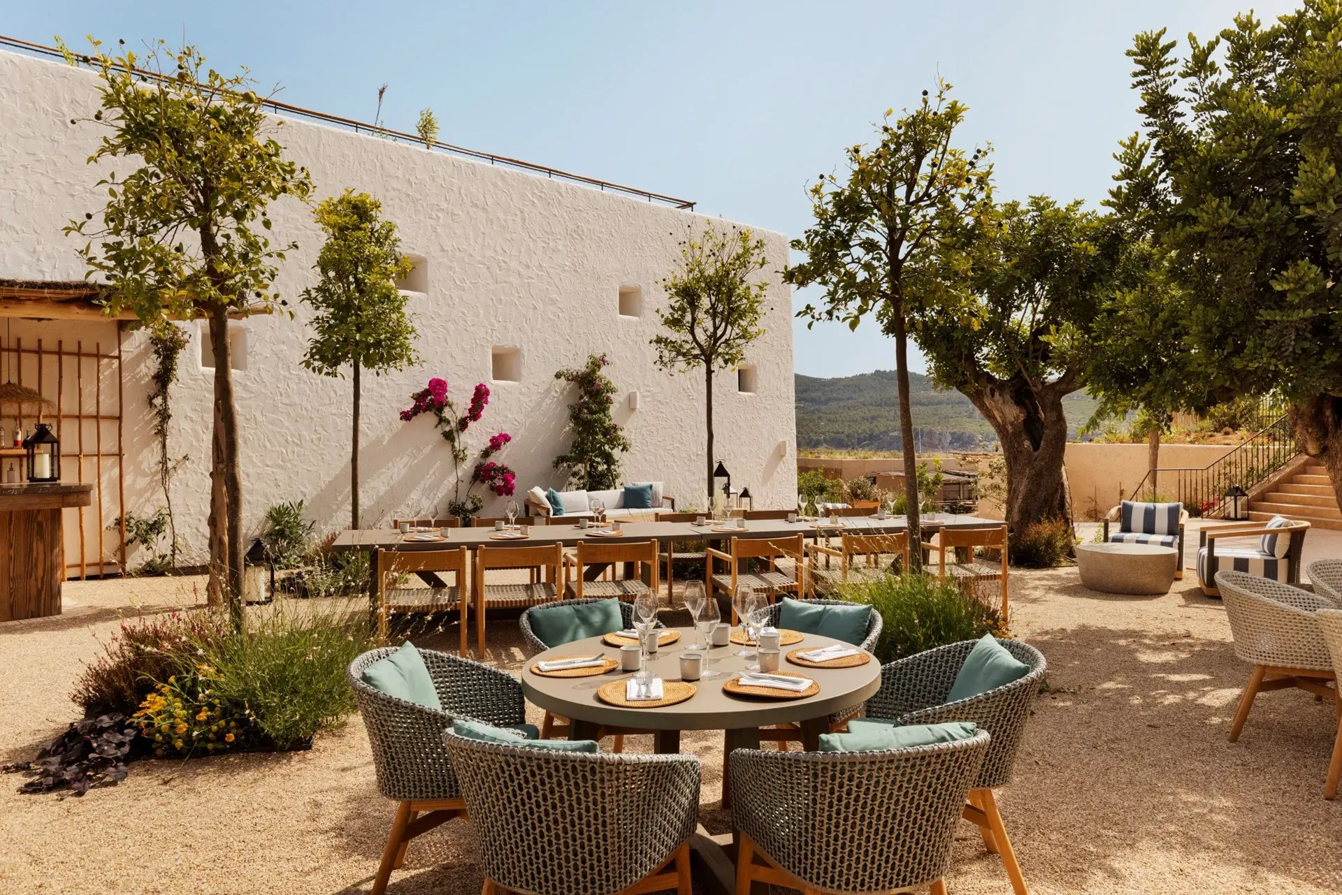 The Orchard, Balearic Islands, Spain — Courtyard dining with woven chairs, round tables, whitewashed walls, mature olive trees, and Mediterranean landscape views.