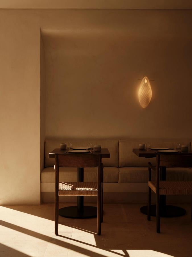 Gundari, Greece — Minimalist dining nook with woven pendant light, wooden chairs, warm plaster walls, and soft natural daylight casting geometric shadows.