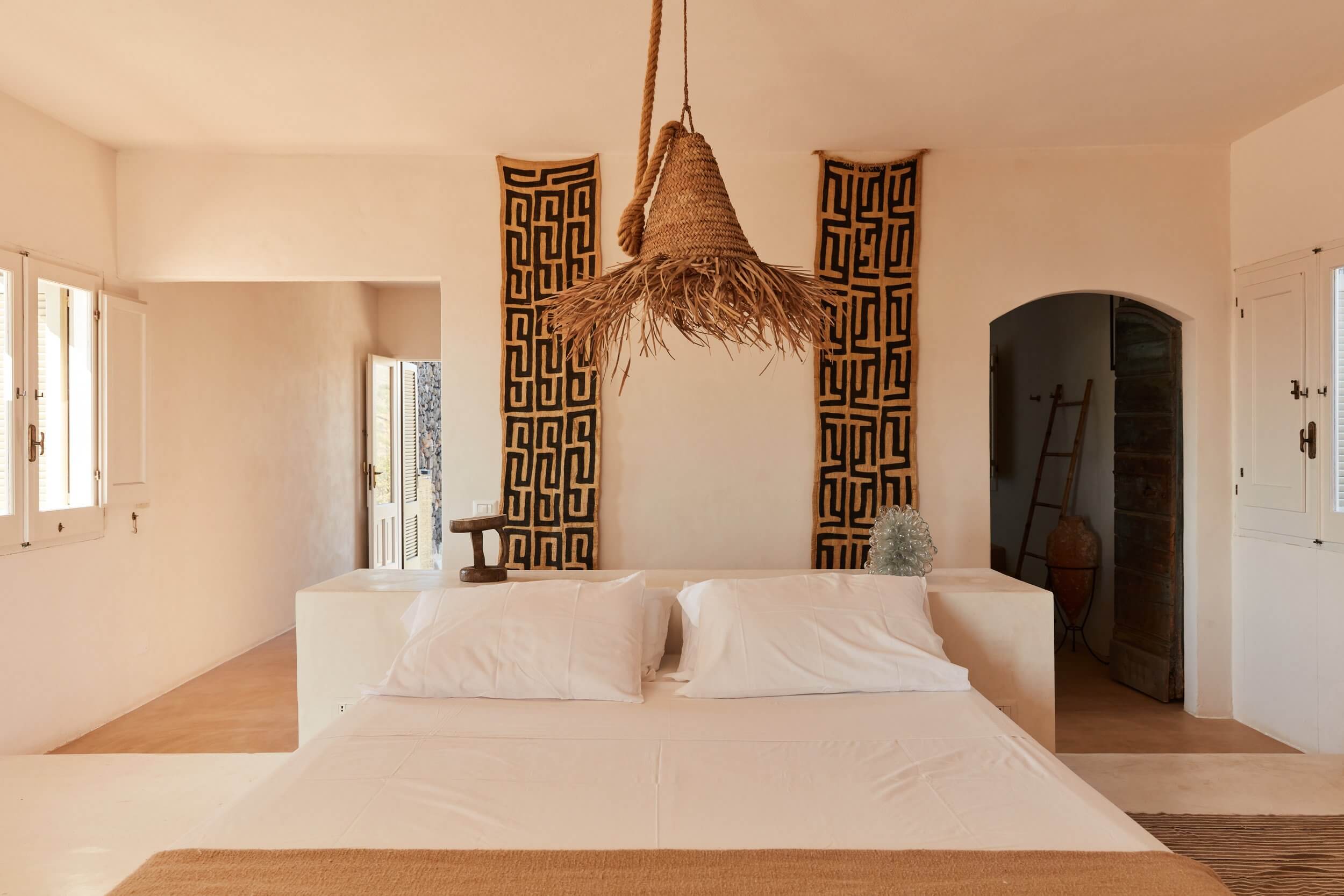 Parco Dei Sesi, Europe — Minimalist bedroom with woven pendant light, geometric wall panels, cream linens, and arched doorway opening to terracotta flooring.