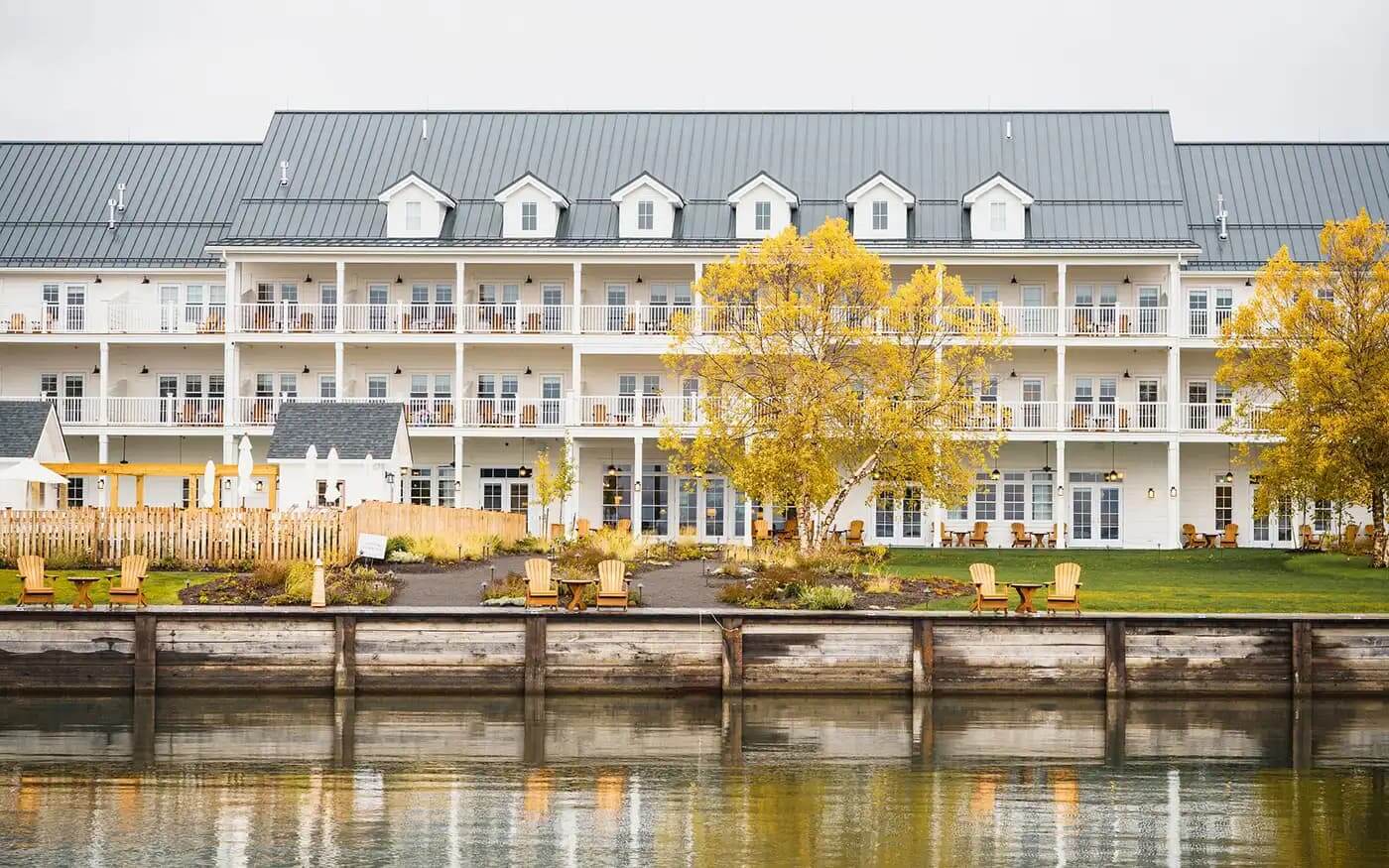 The Lake House, North America — Waterfront luxury hotel with white clapboard facade, metal roof, dormers, and tiered balconies overlooking calm water at golden hour.