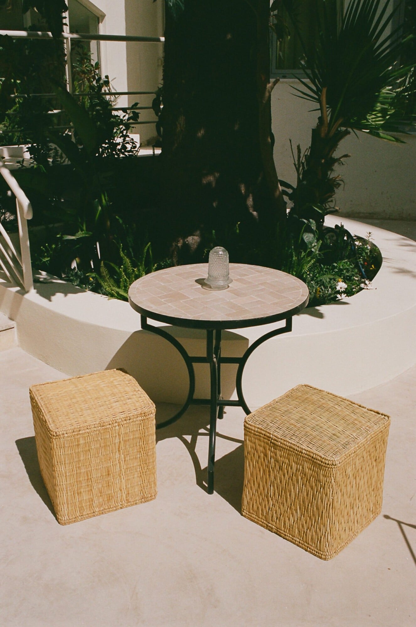 Apollo Palm, Europe — Minimalist courtyard seating with woven natural fiber poufs, round marble-top table, lush green plants, and bright Mediterranean light.
