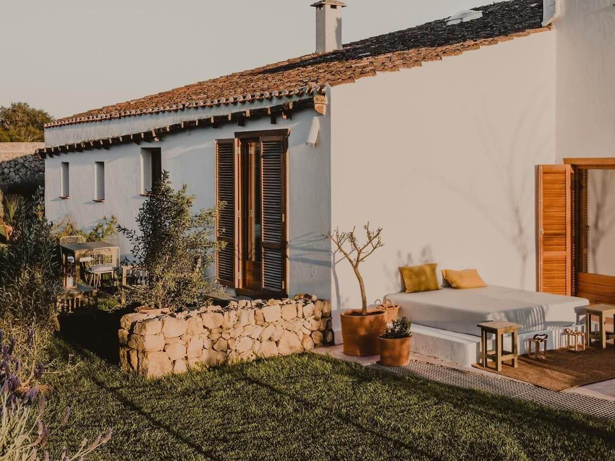 Amagatay, Balearic Islands — Whitewashed Mediterranean villa with terracotta roof, stone walls, wooden shutters, and minimalist outdoor lounge at golden hour.