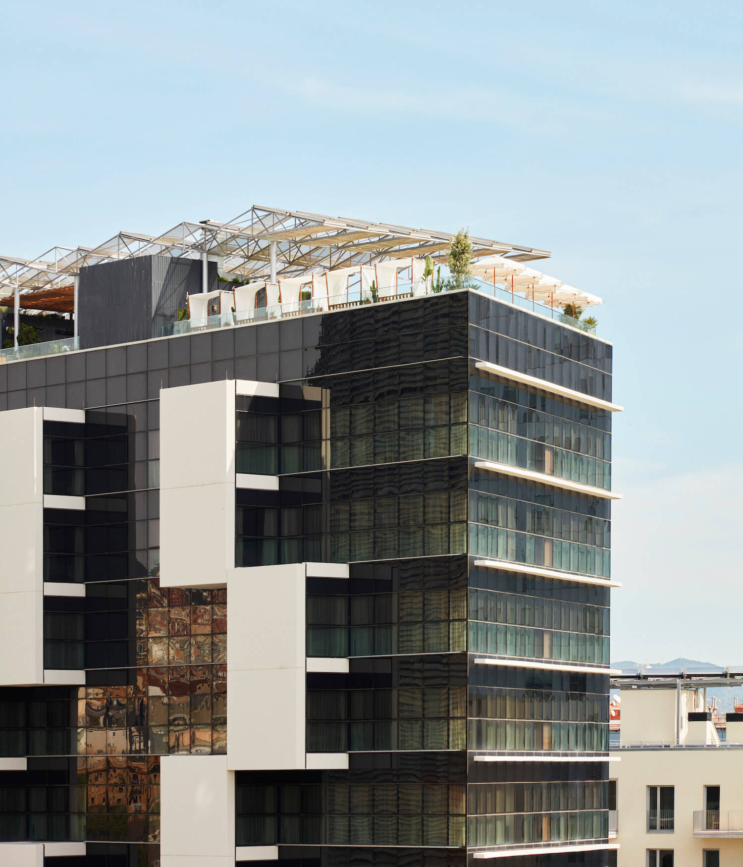 The Hoxton Poblenou, Barcelona, Spain — Contemporary mixed-material facade with black corrugated metal, white stone, and copper panels beneath sculptural timber roofscape overlooking industrial waterfront.