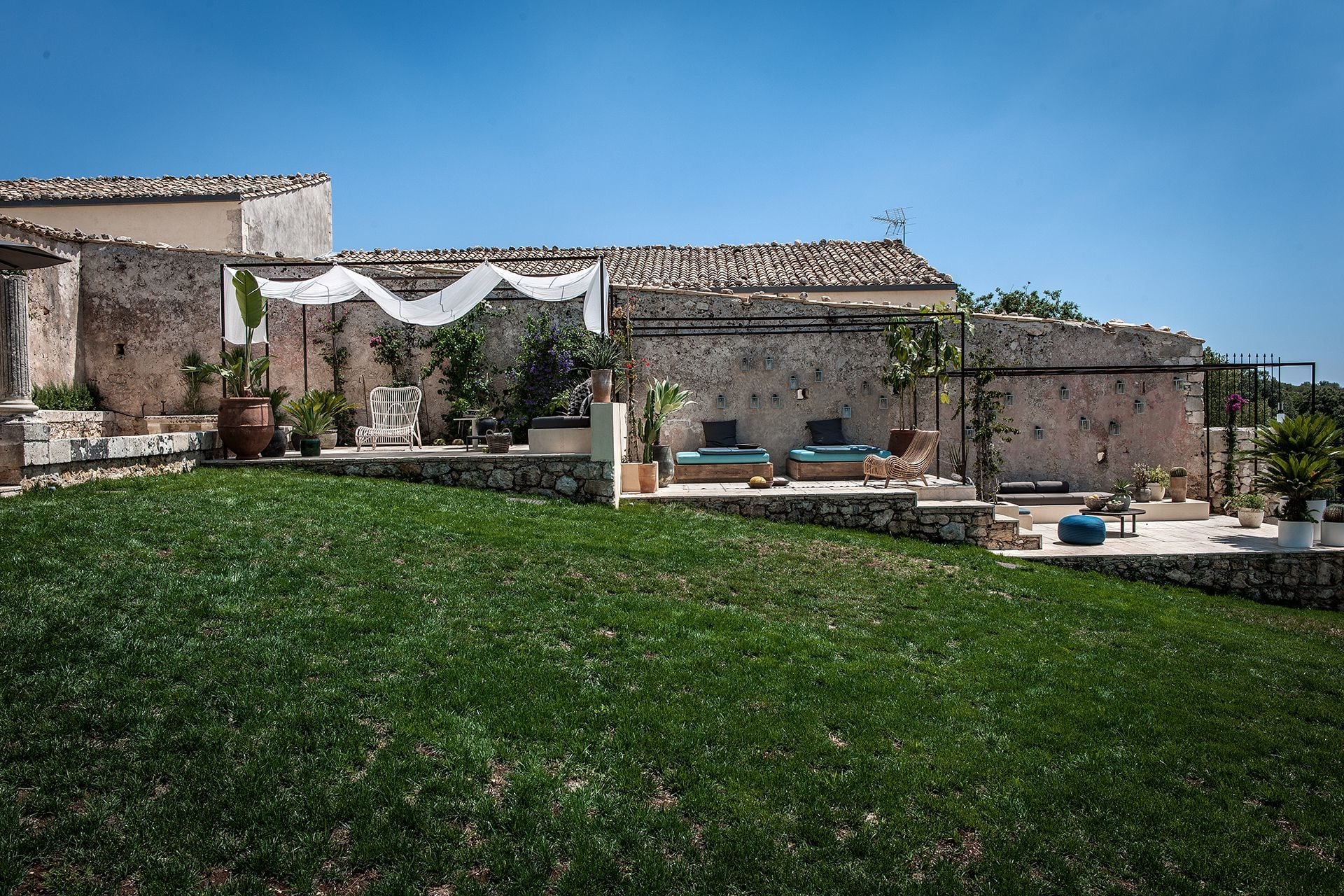 Dimora delle Balze, Europe — Tuscan stone villa courtyard with manicured lawn, terraced lounge areas, white draped canopy, and ochre-tiled roofline under clear sky.