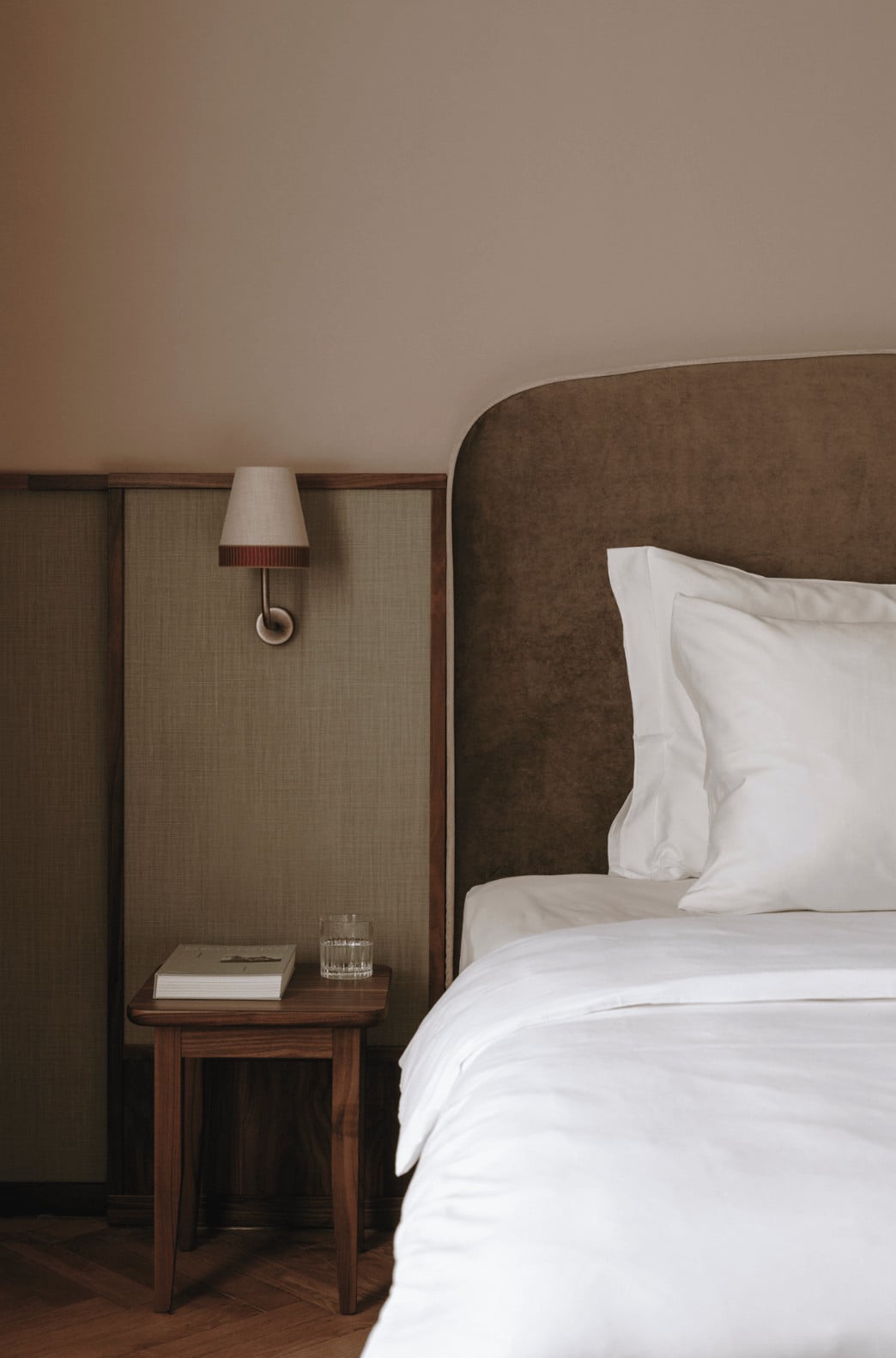 Parkhotel Mondschein, Europe — Brown upholstered headboard with white pillows beside wooden nightstand, minimalist sconce lamp, neutral textured wall paneling, warm hotel bedroom interior.