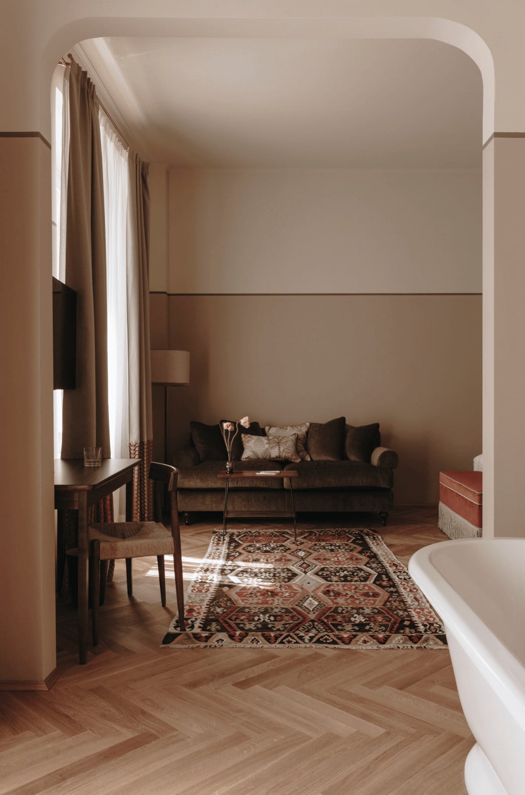 Parkhotel Mondschein, Europe — Spa suite with herringbone wood flooring, taupe walls, dark sofa, geometric kilim rug, arched doorway, natural light from draped windows, freestanding bathtub.