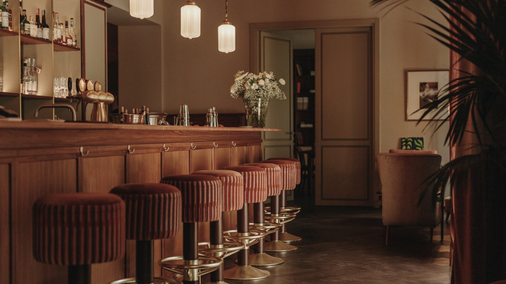 Parkhotel Mondschein, Europe — Warm wood bar counter with burgundy ribbed bar stools, pendant lighting, fresh flowers, and mid-century modern hospitality design.