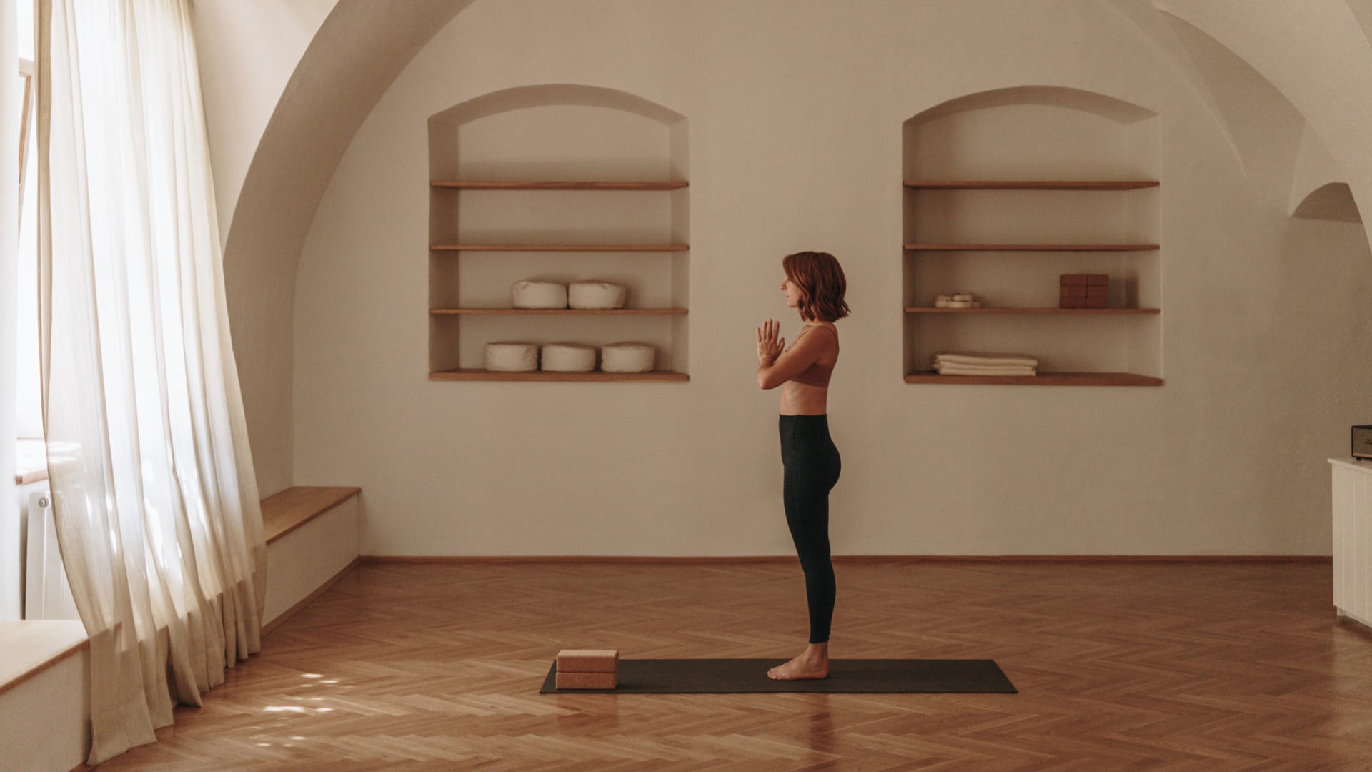 arise Studio, Europe — Woman in yoga pose on mat in minimalist arched room with wood shelving, cream walls, natural light from sheer curtains, warm wood flooring.